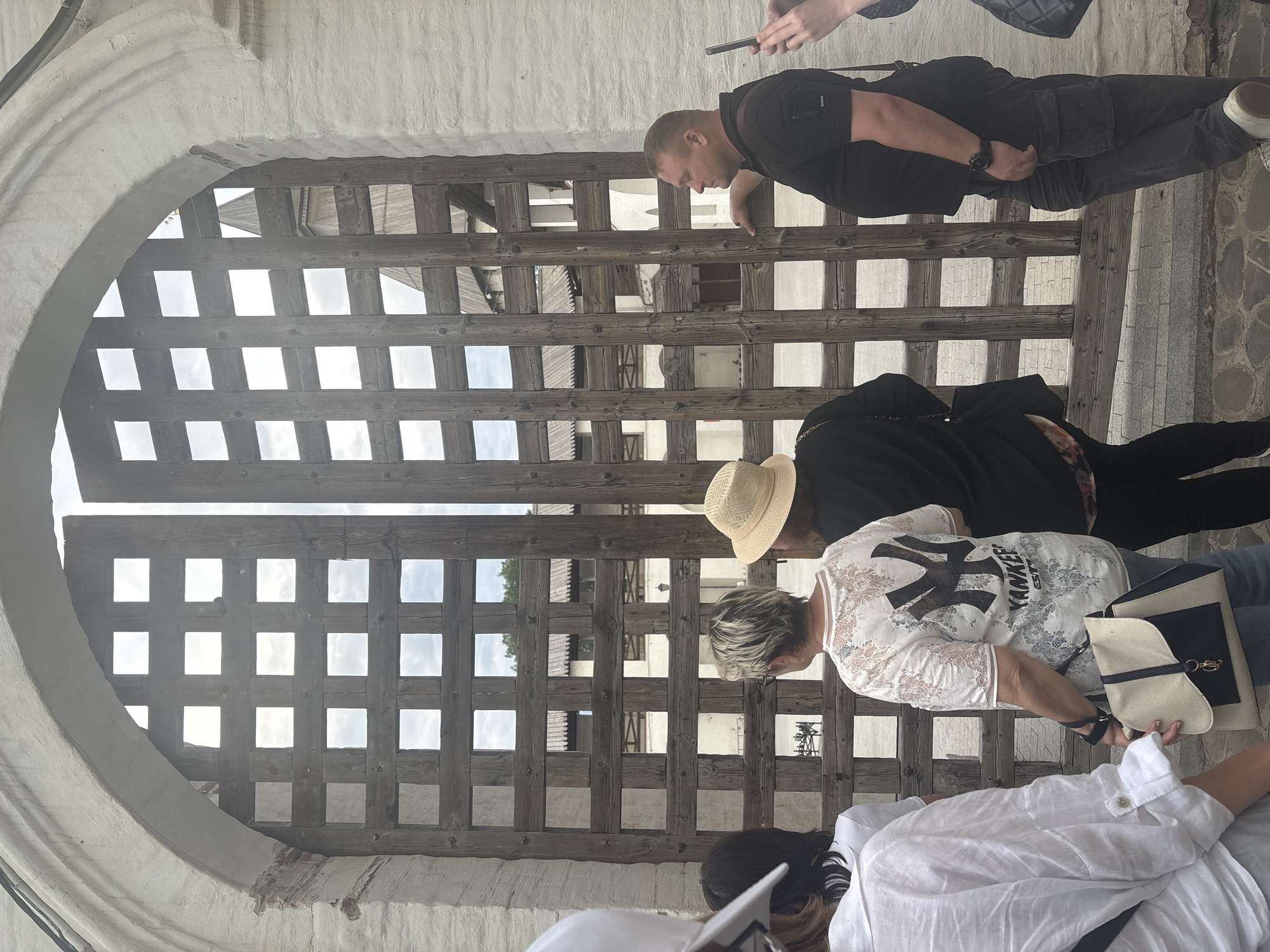 The image depicts an exterior scene at the Astrakhan Kremlin in Astrakhan, Russia, specifically focusing on a large, historical gate. A wide, arched white stone gateway, constructed from textured whitewashed masonry, frames a two-leaf wooden gate. The arch shows signs of age, with some plaster degradation revealing reddish material underneath on the left side, and a dark corrugated pipe running along the top interior curve. The wooden gate is constructed from a robust grid of dark brown, thick wooden planks, forming square openings approximately ten high and ten wide per leaf. The right gate leaf is slightly ajar, creating a narrow vertical gap. Through the gate's grid and the open gap, an interior courtyard or complex of the Kremlin is visible. Distinguishable features inside include white stuccoed buildings, tiered wooden rooflines or structures, and a prominent pointed wooden spire or turret of a building. The sky, showing light clouds, is visible through the upper openings. In the foreground, a group of five to six individuals are positioned, appearing to observe the interior view through the gate. 1. On the far left, a person is partially visible from behind, wearing a white long-sleeved shirt with rolled sleeves and a white cap. 2. Next to them, a person with dark hair is seen from behind, wearing a dark top. 3. In the center-left, a female subject with short, light-colored hair is viewed from behind, wearing a white short-sleeved top featuring a lace pattern and a prominent black "NY YANKEES" logo on the back. She carries a two-tone (beige and dark blue) shoulder bag with a gold-colored clasp. 4. To her right, a female subject is seen from behind, wearing a straw hat and a dark top. 5. On the right side of the frame, a male subject is shown in profile, looking towards the interior. He wears a black short-sleeved polo shirt with a patch on the left sleeve, dark cargo-style pants, and a dark wristwatch on his left wrist. His left arm is casually resting on one of the wooden horizontal beams of the gate. 6. On the far right, two hands are partially visible, one holding a dark-colored smartphone vertically, suggesting another individual is present or interacting with the phone. The ground in the immediate foreground consists of irregular, dark-toned paving stones. The scene is illuminated by natural daylight, indicating an outdoor setting during the day, possibly under an overcast