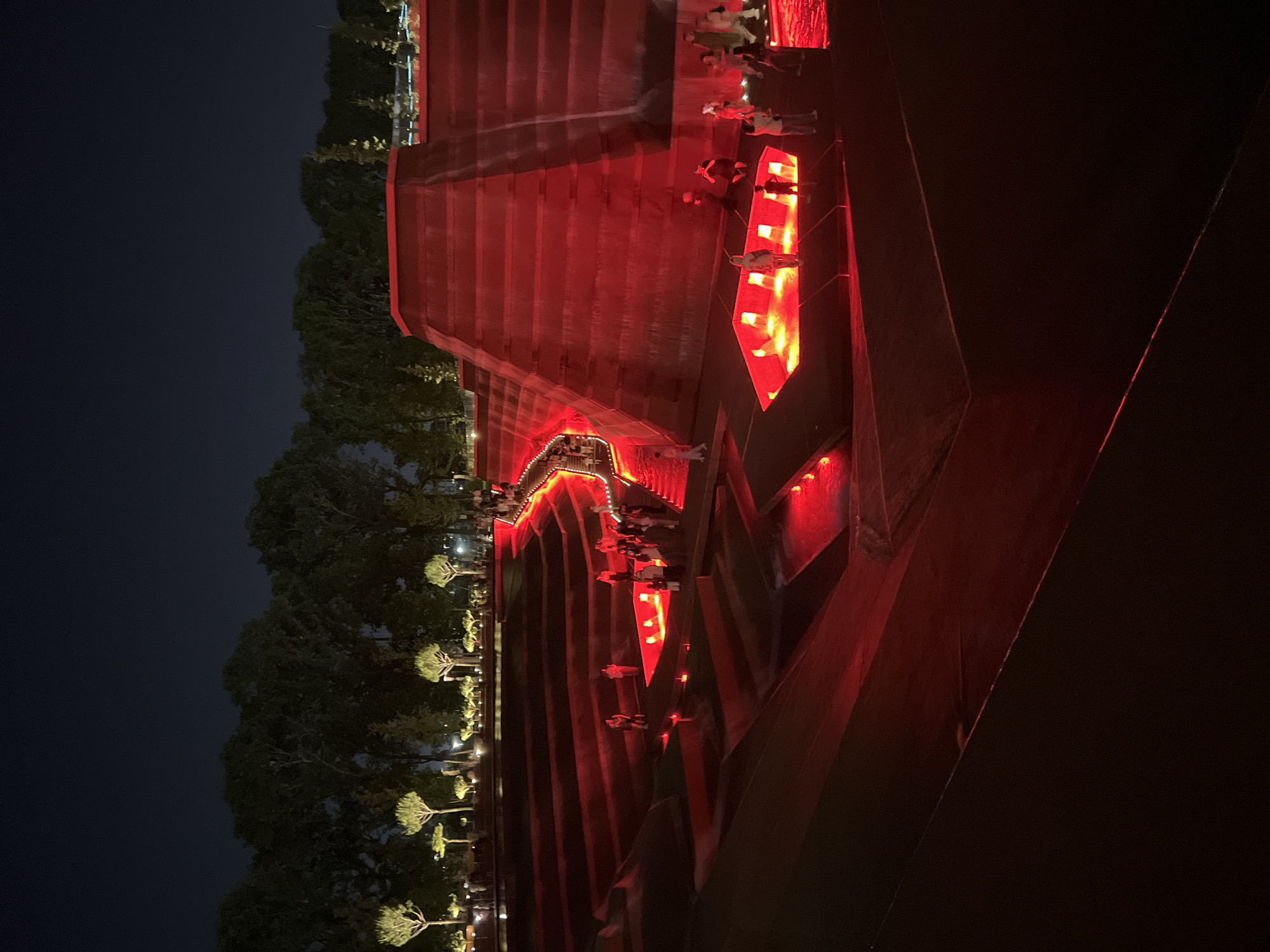 This image depicts a nighttime view of the "Triumphal Arch" park in Krasnodar, Russia. The park features a large, multi-tiered structure with cascading water features, illuminated with vibrant red lights. Several people are visible, walking along the tiered pathways and near the illuminated water features. The surrounding area includes trees and a dark sky. The architecture is modern and geometric, with the red lighting creating a dramatic and atmospheric scene.