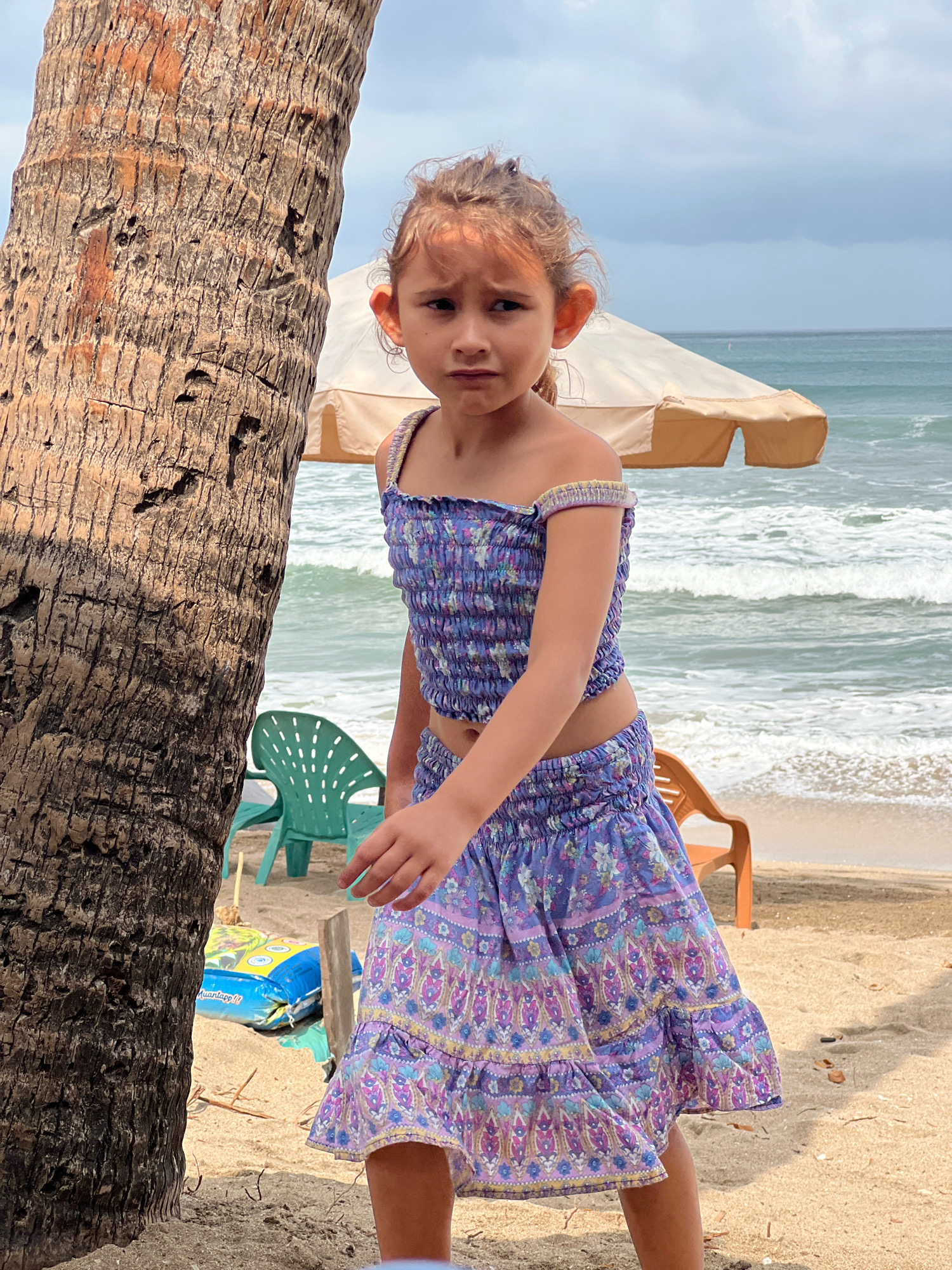 A young girl stands on a sandy beach in Kuta, Indonesia. She is wearing a purple, floral-print smocked crop top and a matching skirt. The girl is positioned to the right of a large palm tree trunk, which takes up the left side of the frame. In the background, the ocean waves gently roll onto the shore under a cloudy sky. An umbrella shades two empty beach chairs, one green and one orange, on the sand. A blue and yellow inflatable object is visible near the base of the palm tree. The girl is looking forward with a neutral expression.