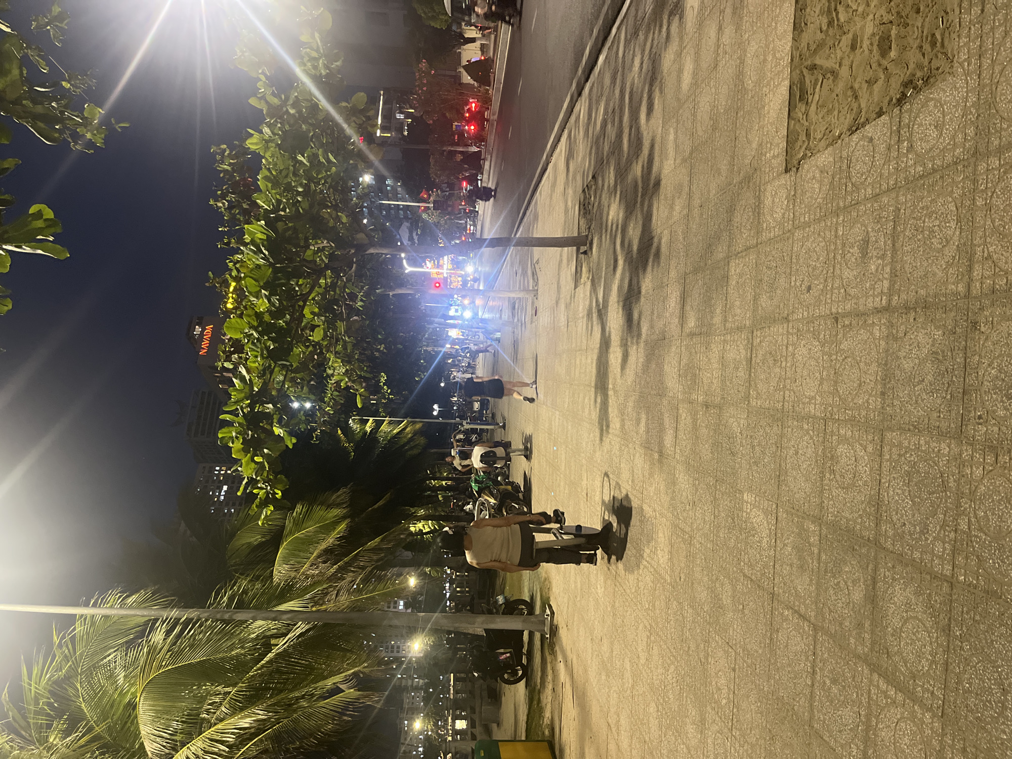 This is a nighttime scene on a brightly lit sidewalk in an unknown city. A street with traffic is visible to the right. The sidewalk is paved with rectangular tiles. Two people, appearing to be conversing, are walking to the right on the sidewalk. Parked motorbikes and palm trees line the left of the sidewalk. There is a tall building with a “Navada” sign in the background. Street lights are visible, causing glare.
