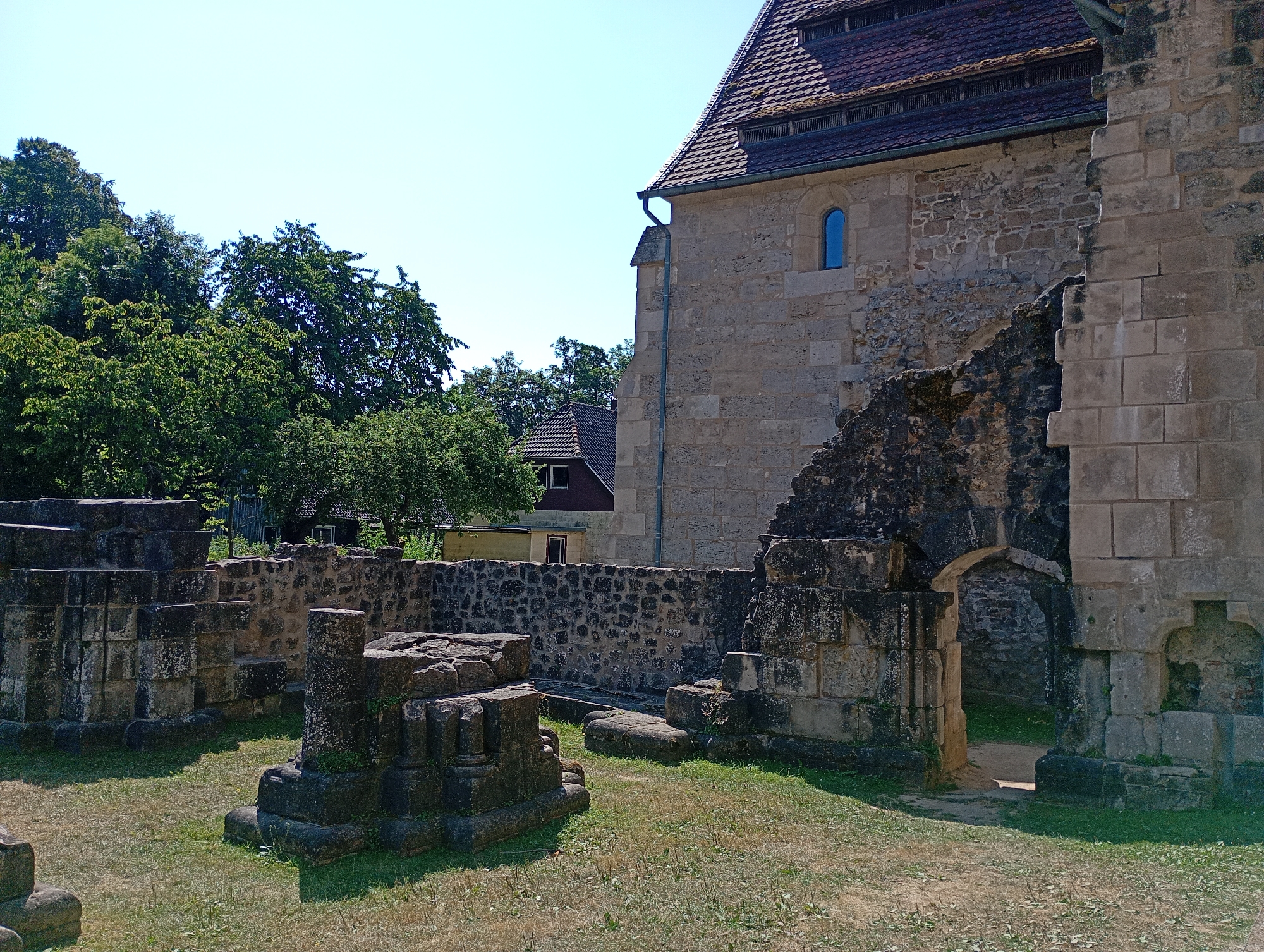 The image shows the exterior of the ruins of the Walkenried Abbey in Walkenried, Germany. In the foreground, there are stone ruins and overgrown grass. The ruins appear to be made of large, rough-hewn stones, with some sections appearing more intact than others. One section has an arched entryway leading into a dark space. In the middle ground, there is a stone wall and further ruins. In the background, there are trees and buildings, including a large building with a tiled roof and a small blue window. The sky is clear and blue.