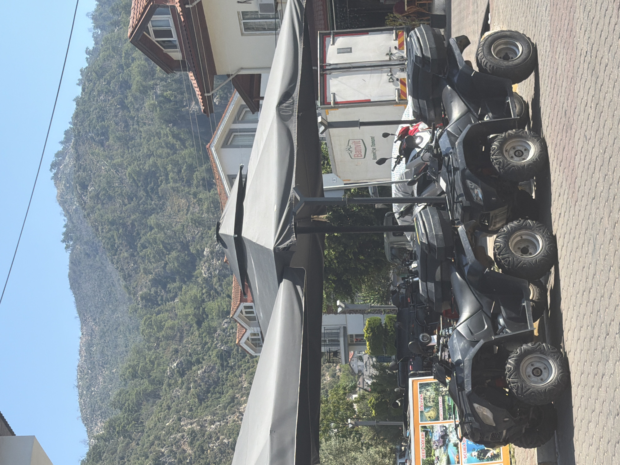 The image depicts a row of parked black ATVs, or quad bikes, on a cobblestone street in Marmaris, Turkey. The ATVs are lined up under a large black umbrella canopy, suggesting they are available for rent or part of a tour operation. In the background, a white refrigerated truck with "Banvit" branding is visible, as well as modern buildings with tiled roofs and an air conditioning unit. Beyond the buildings, a lush green, mountainous landscape under a clear blue sky dominates the scene. One ATV has a poster attached to its front with images of activities like "Jeep Safari," "Dalyan," and "Turtle Beach," further indicating a tourist-oriented location. There are no people visible in the frame.