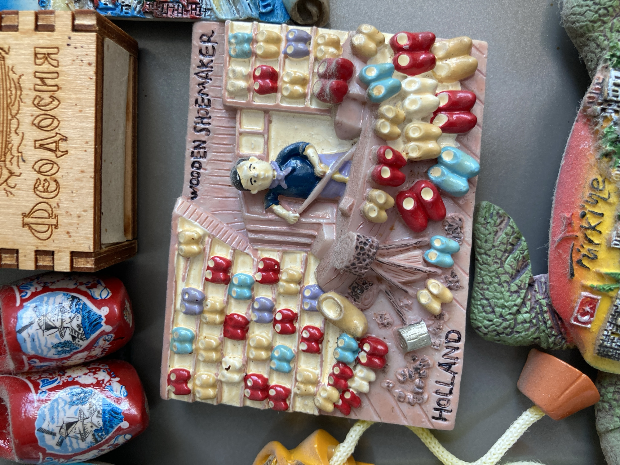 This is a close-up, eye-level shot of a collection of refrigerator magnets. The central magnet depicts a miniature shoemaker's shop with shelves stocked with tiny wooden clogs in various colors, including red, blue, yellow, and purple. A small figure of a shoemaker, dressed in a blue shirt and apron, stands at a workbench holding a tool. The magnet is labeled "WOODEN SHOEMAKER" and "HOLLAND." To the left of this magnet are two red and white Dutch clogs with windmill imagery. Above the shoemaker magnet is a wooden box with Cyrillic lettering that appears to read "Феодосия" (Feodosiya), suggesting a Crimean origin. Below the shoemaker magnet, parts of other magnets are visible, including a green, textured magnet resembling foliage and another magnet with a colorful depiction of buildings and the word "Türkiye." The magnets are placed on a gray, metallic surface.