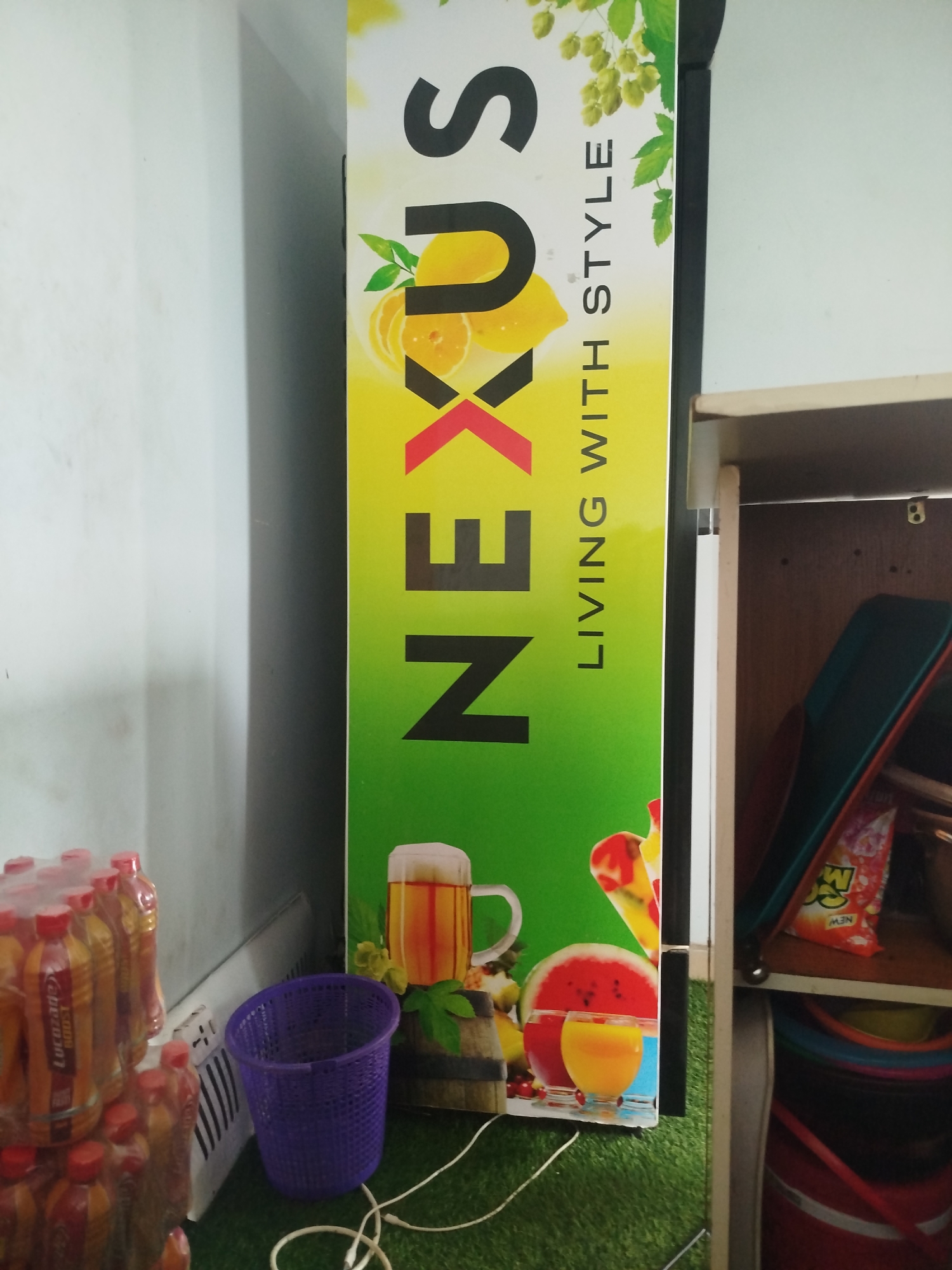 A vertical, full-frame, eye-level shot captures a tall, slim, brightly colored advertisement display standing on a green, artificial turf surface in a room. The advertisement, predominantly yellow and green, features the word "NEXUS" in large, black, bold, sans-serif letters vertically oriented down the center. To the right of the word "NEXUS" is the phrase "LIVING WITH STYLE" also in black text. The top portion of the advertisement shows green foliage and what appear to be hops. Below "NEXUS", there are graphic representations of sliced oranges, a beer mug, a watermelon, and two glasses of juice. The advertisement is positioned against a white wall on the left. To the right of the advertisement, a wooden shelf unit is visible, containing various items, including what appears to be a black bag and several colorful bowls or buckets stacked on top of each other. In the foreground to the left of the advertisement, a stack of plastic bottles, many with orange and yellow labels, is present. A purple, woven basket sits on the green turf between the bottles and the advertisement. A white electrical cord lies on the turf in front of the advertisement. The overall setting appears to be indoors, possibly a shop or a storage area, in Uselu, Nigeria. No people are visible in the image.
