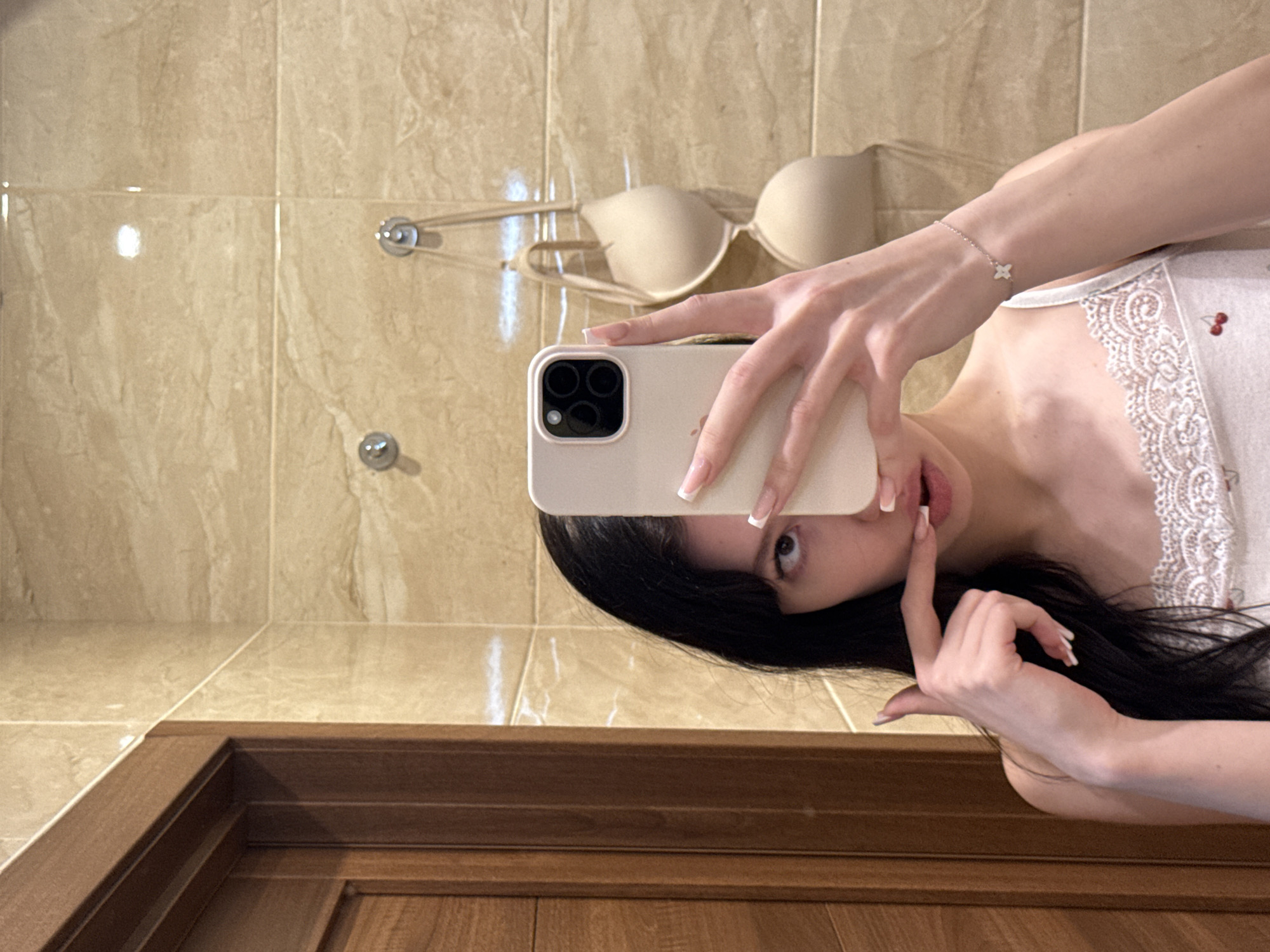A young woman with long, dark hair and bangs is taking a selfie in a bathroom. She is wearing a white camisole with lace trim and a cherry print. Her left hand is holding a smartphone, and her right index finger is touching her lips. In the background, a beige bra hangs from hooks on a tiled wall. A wooden door frame is visible to the left of the woman. The lighting is bright, creating reflections on the tiled surfaces.