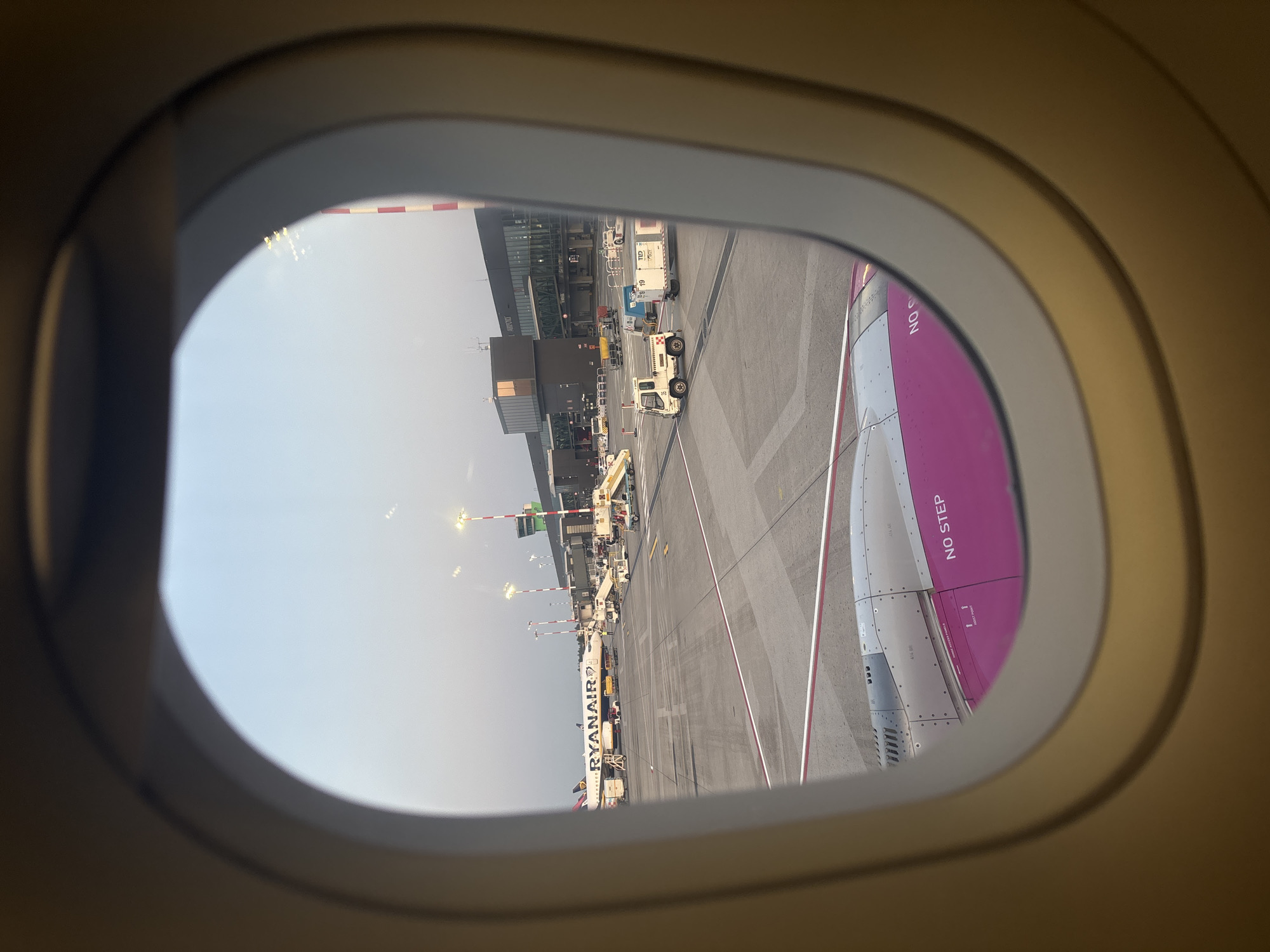 This is an external view from an airplane window at Orio al Serio International Airport (BGY) in Orio al Serio, Italy. The image shows an airport tarmac with several aircraft and ground support equipment. A Ryanair airplane is visible on the left, parked at a gate. Various service vehicles, including baggage carts and a pushback tractor, are positioned around the aircraft. In the background, the airport terminal building and a red and white striped control tower are prominent. The sky is clear and appears to be late afternoon or early evening, casting a soft light on the scene. Part of the wing and engine of the airplane from which the photo is taken is visible in the foreground, displaying the "NO STEP" warning.
