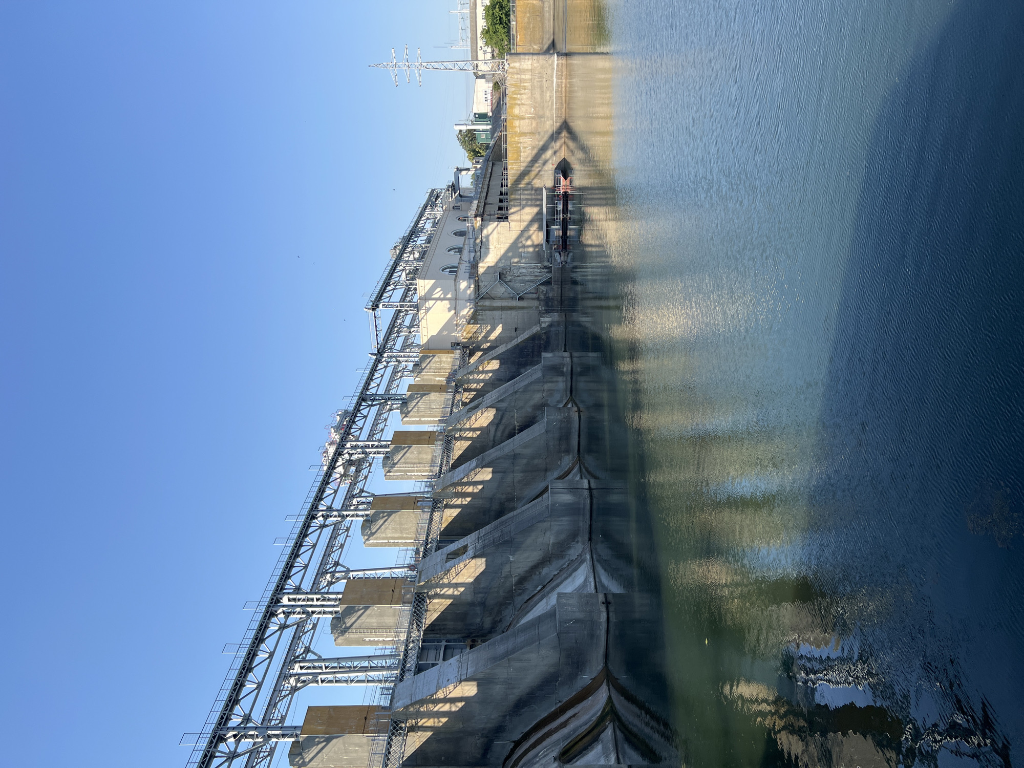 A wide shot captures the impressive scale of the N. B. N. dam, a concrete structure spanning across a tranquil body of water. The dam's angled spillways, cast in shadow by the morning sun, descend towards the water's surface, reflecting the stern geometry of the architecture. A network of overhead metal walkways and supporting structures extends across the top of the dam, hinting at the complex machinery and operations within. The clear blue sky above provides a stark contrast to the massive, man-made edifice. In the foreground, the water is calm, with subtle ripples distorting the reflections of the dam. A small, flat-bottomed boat is moored to a platform near the dam's central section, suggesting human presence and activity in this industrial landscape. The overall scene is one of immense power and engineering prowess, set against the natural backdrop of a clear sky and calm water. The specific location of N. B. N. dam in Ustia, Moldova is indicated.