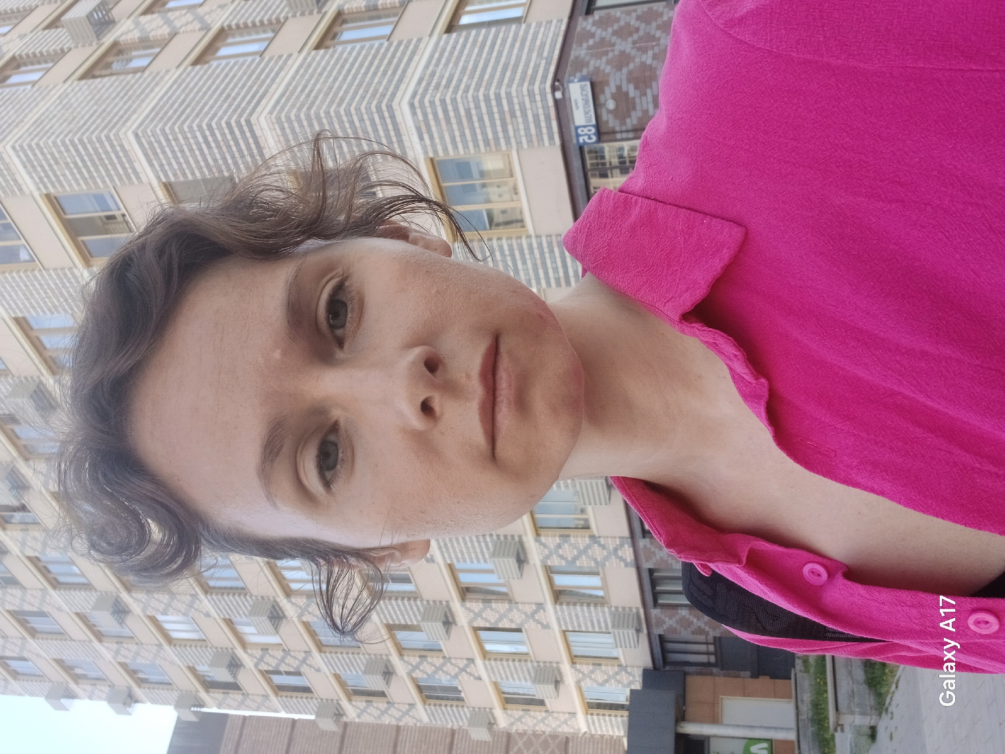 A fair-skinned woman with curly brown hair wears a vibrant pink button-down shirt. She is standing in front of a tall, modern building with rows of windows. A street sign with the number "28" is visible on the building. The photo appears to have been taken in Yekaterinburg, Russia, as indicated by the street address plaque with the number "28" visible on the side of the building behind the woman.