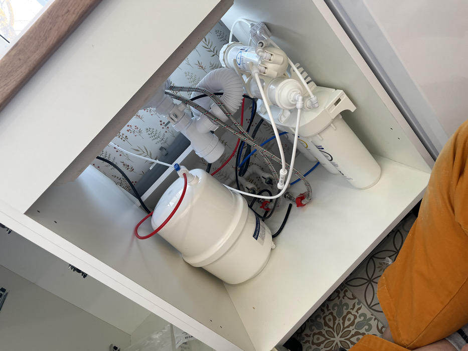 The image displays the interior of a white cabinet, likely beneath a sink, containing a water filtration system and plumbing components. A white, cylindrical pressure tank for reverse osmosis is positioned on the left, connected by a red tube at its top. To the right, multiple white water filter housings are interconnected by blue, red, white, and black plastic tubing and various fittings. A white plastic P-trap and a flexible white corrugated drain hose are visible in the upper right, along with two braided stainless steel water supply lines connected to valves. The cabinet's rear wall is covered with light wallpaper featuring a repeating botanical pattern of green leaves and small red elements. A wooden countertop edge is partially visible at the top right. In the lower-left quadrant, the legs of an individual wearing orange pants are visible. The floor outside the cabinet features patterned tiles. The location of Нарынка, Russia, cannot be confirmed from the visual information present in the image.