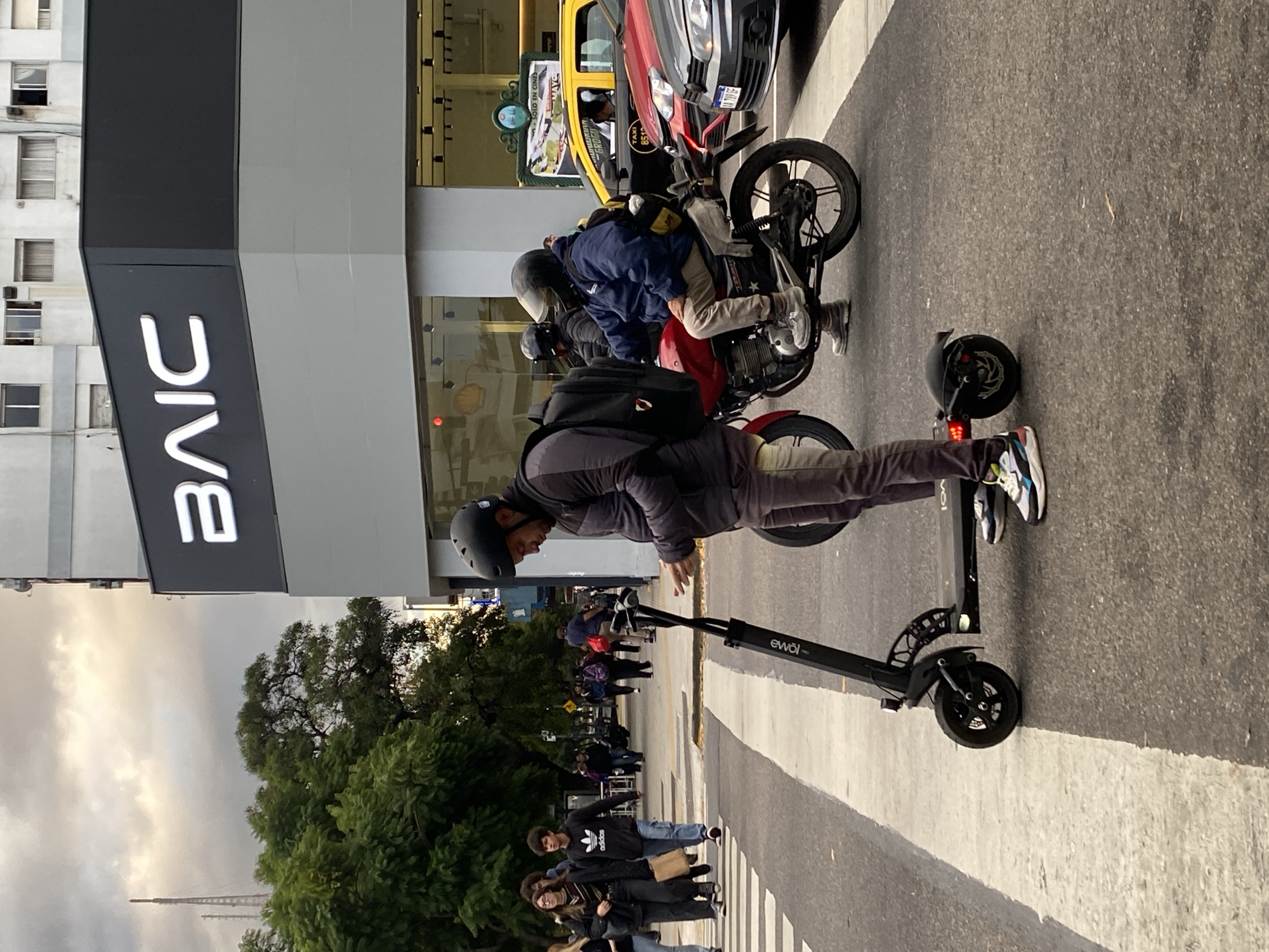 The image shows a man on an electric scooter, positioned on a zebra crossing in what appears to be an urban environment. The scooter has "eWOi" visible on its side. He is wearing a helmet and casual clothing, including sneakers. Behind him are other pedestrians using the crosswalk. Further back, there are two motorcyclists, one seated on his bike. Taxis and other vehicles are also visible on the road. A building with the letters "BAIC" prominently displayed is on the left side of the frame. The presence of the BAIC sign and architectural styles strongly suggest the location is in Buenos Aires, Argentina. There are trees and other buildings visible, including what appears to be the Buenos Aires Obelisco in the distance, confirming the city. The sky is overcast, and the overall lighting suggests either early morning or late afternoon.