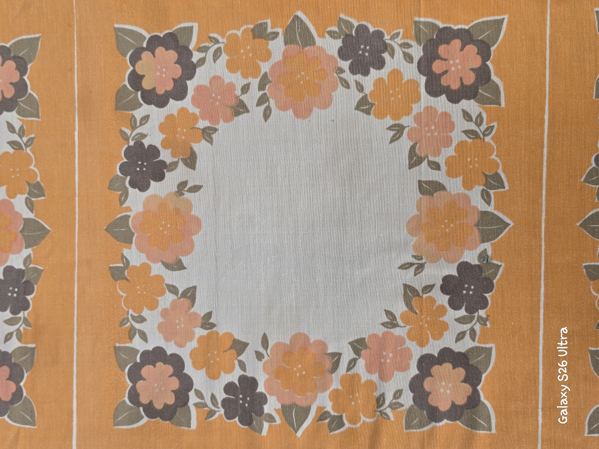 This is a close-up, top-down view of a patterned fabric, likely a tablecloth or placemat. The pattern consists of stylized flowers with five petals and small leaves, arranged in a decorative border around a central light-colored area. The flowers are depicted in shades of orange, pink, and dark brown, with brown leaves. The background fabric has a textured appearance. The outer edges of the fabric are a solid, bright orange with a thin white vertical stripe running down the right side. The overall impression is of a retro or vintage aesthetic. There is no discernible activity or event taking place, nor are there any people or discernible emotions conveyed by the image. The location context of Plovdiv, Bulgaria, is provided but not visually evident in the image itself. Visible text on the right side reads "Galaxy S26 Ultra".