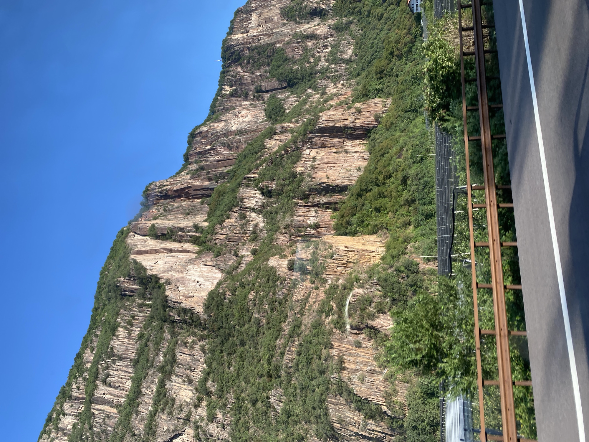 The image shows a steep, rocky mountain face covered in green vegetation, with a clear blue sky above. In the foreground, there is a road with a white line down the middle, bordered by a rusty metal guardrail. Behind the guardrail, there is an area with rows of what appear to be solar panels or agricultural netting, interspersed with trees and shrubs. A residential building is visible in the distance on the right side of the frame. The overall scene suggests a natural landscape adjacent to human infrastructure, possibly on the outskirts of a town or city. The location is identified as Vadena - Pfatten, Italy.