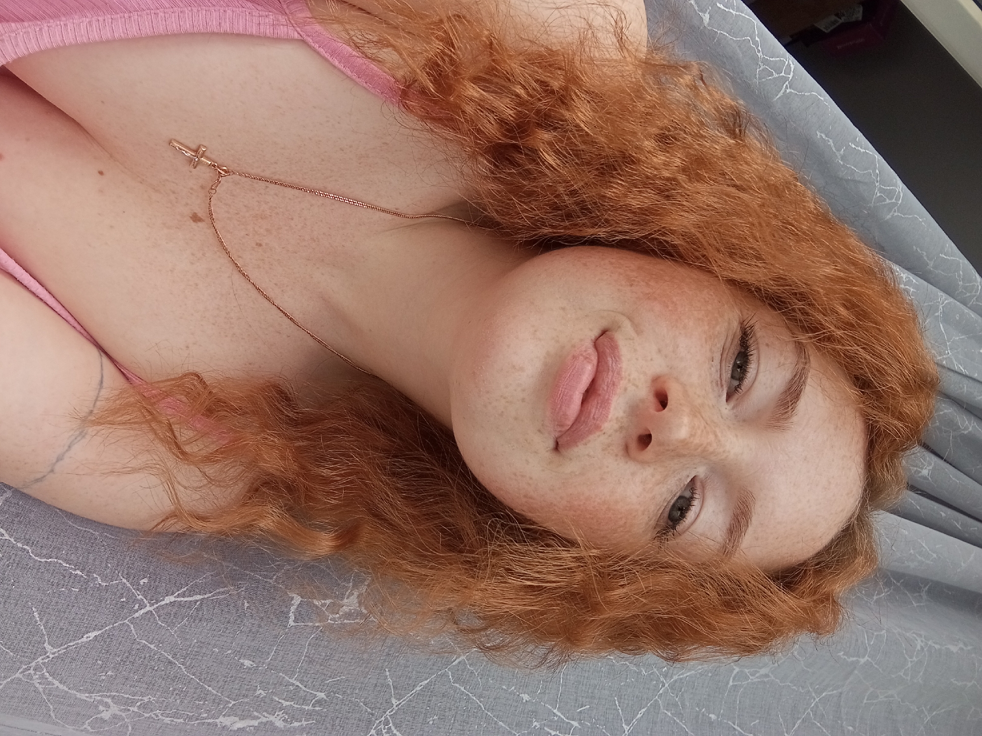 A close-up, high-angle shot features a young woman with red, curly hair lying on a gray surface with a white marble-like pattern. She wears a light pink top. A thin, gold chain necklace with a cross pendant rests on her chest. The woman's face is tilted, and she is sticking out her tongue slightly. She has freckles. The location is unknown and there is no basis to assume that this is Yurgamysh, Russia.