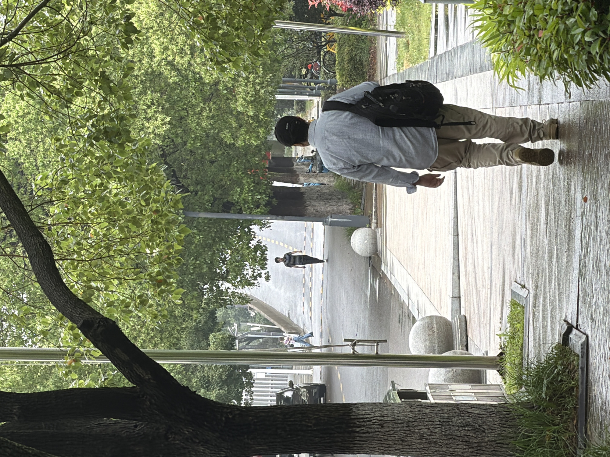 A man wearing a light gray long-sleeved shirt, khaki pants, a black backpack, and a black baseball cap is walking along a wet sidewalk. The sidewalk is lined with foliage on the right and a tree on the left. In the distance, another person is walking in the opposite direction on the street. A car is parked along the road. The setting appears to be a street in Jiangning District, China, based on the urban landscape and foliage.