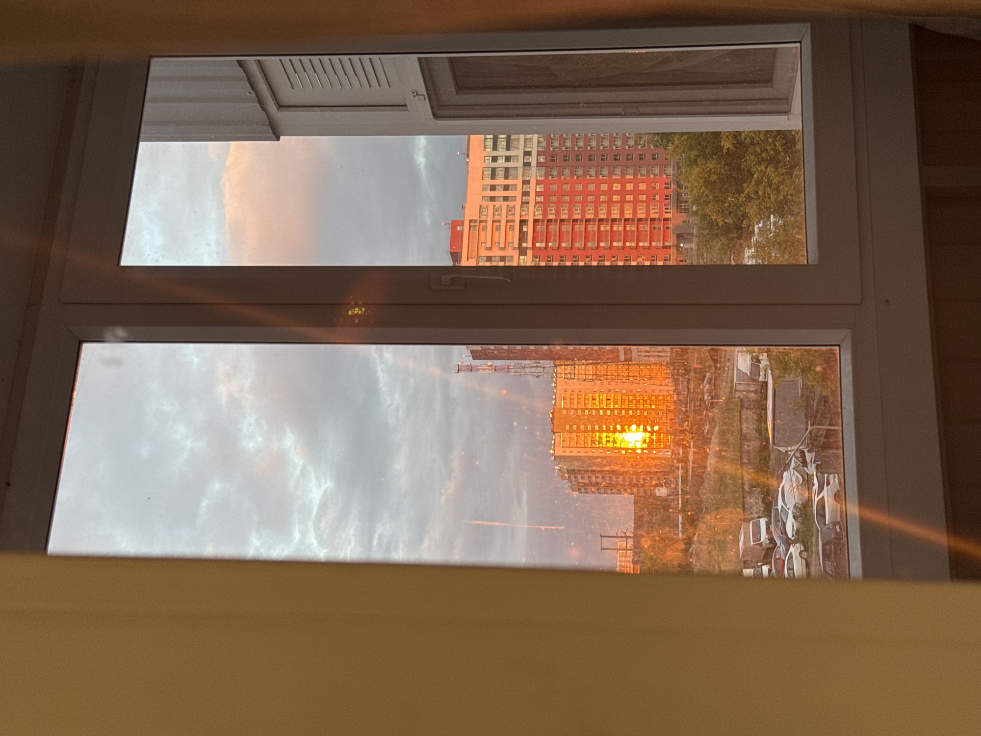 The image captures an urban landscape in Кондратово, Russia, viewed through a multi-pane white window from an interior vantage point. The sky is largely overcast with grey clouds, displaying subtle pink and orange hues characteristic of dawn or dusk. A single, thin white contrail is visible vertically in the upper-left sky.
In the midground, a prominent multi-story building on the left is intensely illuminated by direct sunlight, causing its facade to glow a vivid orange. To its right, another large multi-story building, primarily red and white, is also bathed in the warm light. A tall metal communication tower stands between these structures. Below the buildings, an unpaved lot on the left serves as a parking area for several vehicles, including white vans, dark cars, and a truck. Green trees and dense foliage are visible beneath the red and white building on the right.
The white window frame bisects the view. The window panes exhibit numerous specks, possibly dust or water spots, and prominent light streaks from sun glare. Yellowish-tan wall sections frame the window on both sides, with a portion of a white louvered shutter visible on the far right within the frame. No people are present in the scene.
