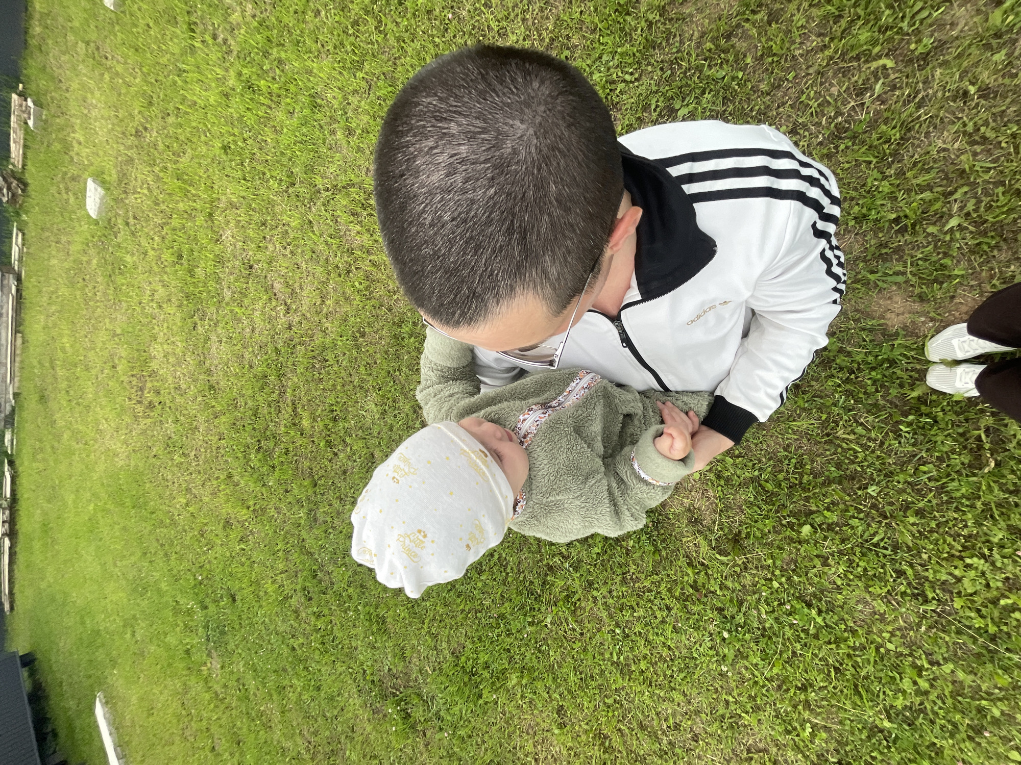 An adult, wearing glasses and a white Adidas tracksuit with black stripes on the sleeves, is holding a baby who is wearing a green fluffy jacket and a white bonnet. The baby's bonnet has gold lettering that reads "Little Prince" and other small golden designs. They are sitting on a patch of green grass, with some white gravestones visible in the background. The image is taken from a high angle, looking down at the subjects. The overall setting appears to be a graveyard on a cloudy day. The adult's facial expression is obscured by their proximity to the baby, but the scene conveys a sense of gentle care and connection between the two individuals.