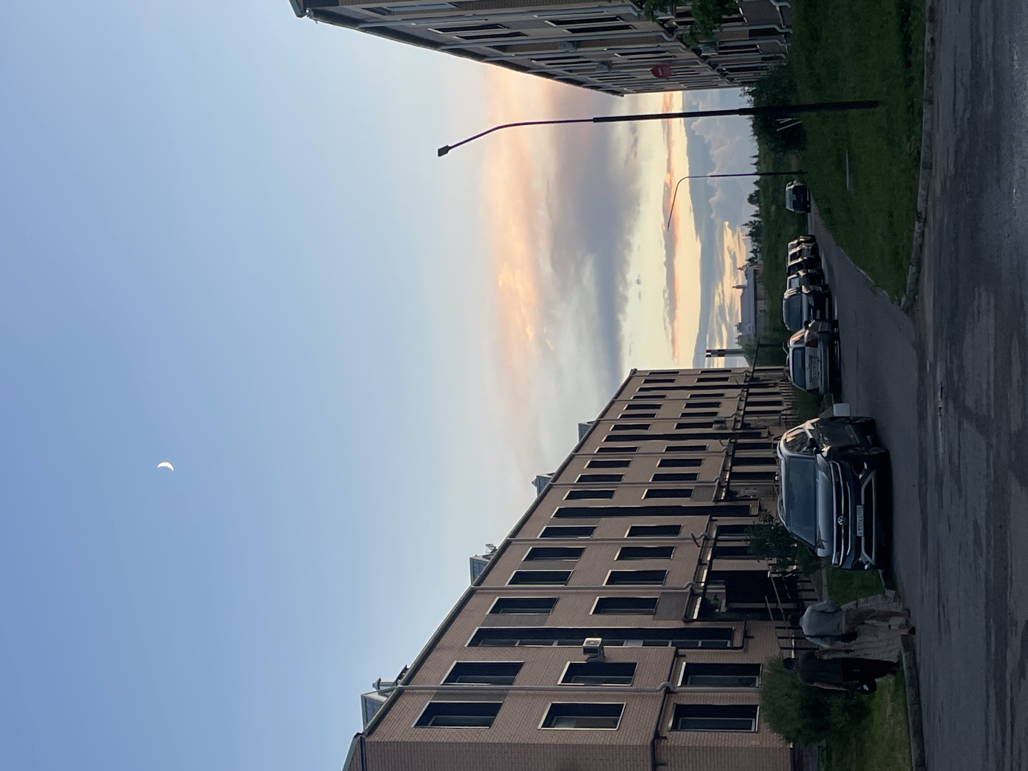 The image depicts a street scene in Zverevo, Russia, under a pale blue sky with a crescent moon visible. On the left, a long, multi-story brick building with numerous windows runs along the street. Two individuals, a man in a dark t-shirt and another in a light-colored shirt and light-colored pants, walk away from the camera along the sidewalk. In front of them, a dark-colored SUV is parked. Several other cars are parked in a line further down the street on the right. The right side of the scene is dominated by another similar multi-story brick building, with a street lamp in the foreground. The sky transitions to hues of pink and orange near the horizon, indicating either sunrise or sunset, with scattered clouds. Green grass and some vegetation are present alongside the buildings and the street.