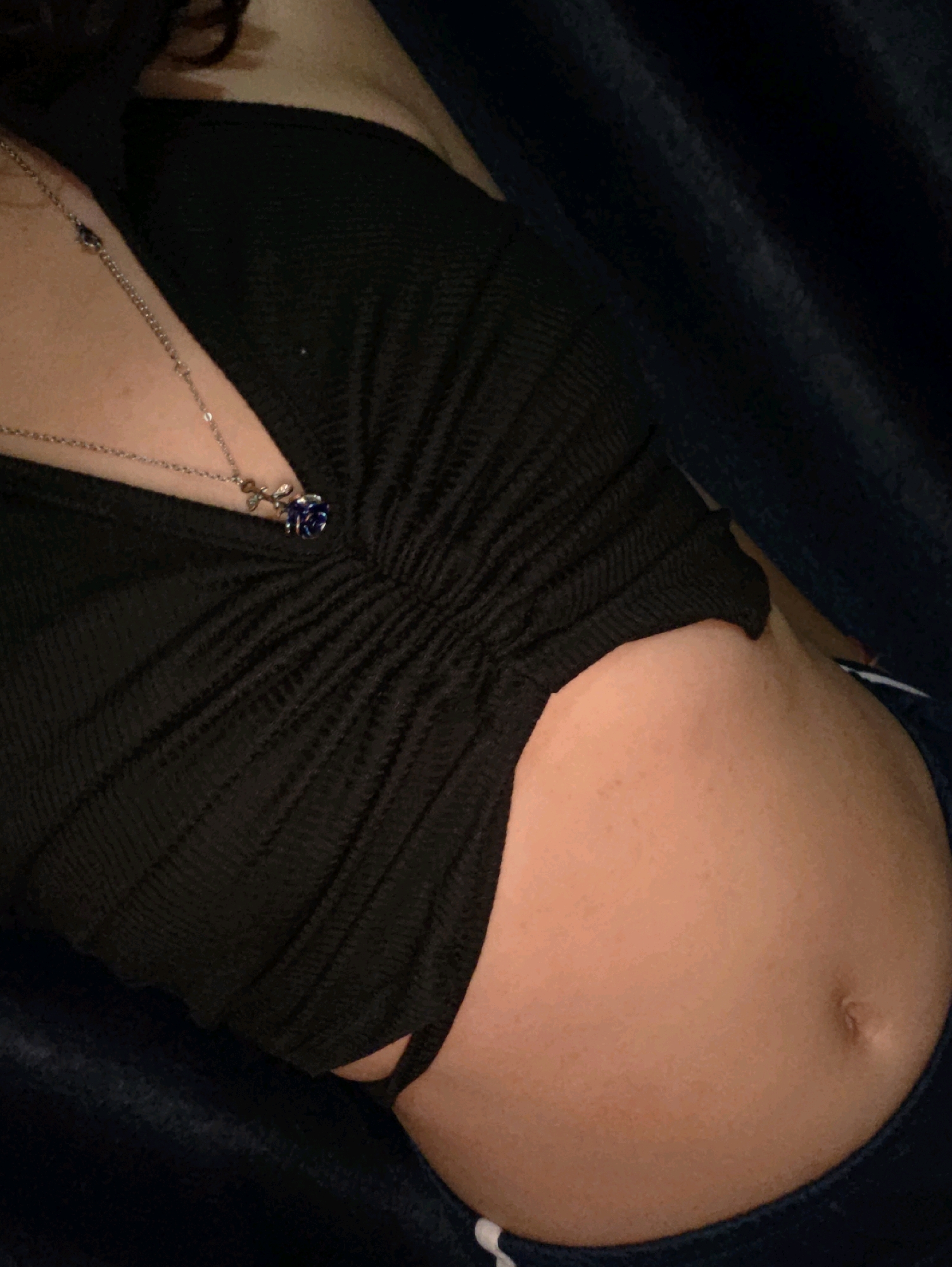 The image shows a close-up of a person's torso, with a focus on their stomach and chest area. They are wearing a black, ruched top and a necklace with a blue pendant shaped like a rose. The background is dark and appears to be fabric, possibly a blanket or clothing. The person is lying down. The image does not provide any information or context related to the city of Phan Thiết, Vietnam.