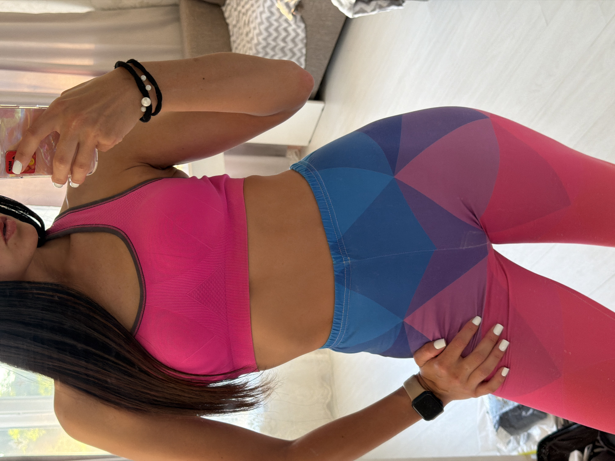 A medium shot captures a woman from the chest down, standing in what appears to be a bedroom. She is wearing a vibrant pink sports bra and colorful leggings with a geometric pattern of pink and blue. Her left arm is resting on her hip, and her right hand is holding a smartphone as if taking a mirror selfie. A watch is visible on her left wrist, and a bracelet adorns her right wrist. The background reveals a bed with a patterned duvet and curtains, suggesting an indoor residential setting. The lighting is bright, possibly from natural light sources. The scene is devoid of any specific Sochi landmarks or recognizable city elements, making it impossible to pinpoint the exact location within Sochi, Russia, based solely on the visual information provided.