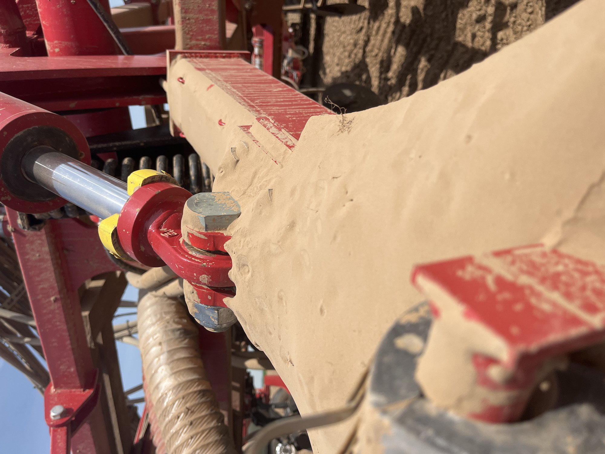 The image shows a close-up of a piece of heavy machinery, possibly agricultural equipment, covered in a thick layer of tan-colored dust or dirt. The machinery appears to be red with some silver and yellow components. A hydraulic cylinder is visible, along with nuts and bolts connecting different parts of the machinery. The environment seems dry and dusty, suggesting an arid or agricultural setting. The location is specified as Unknown, Unknown.