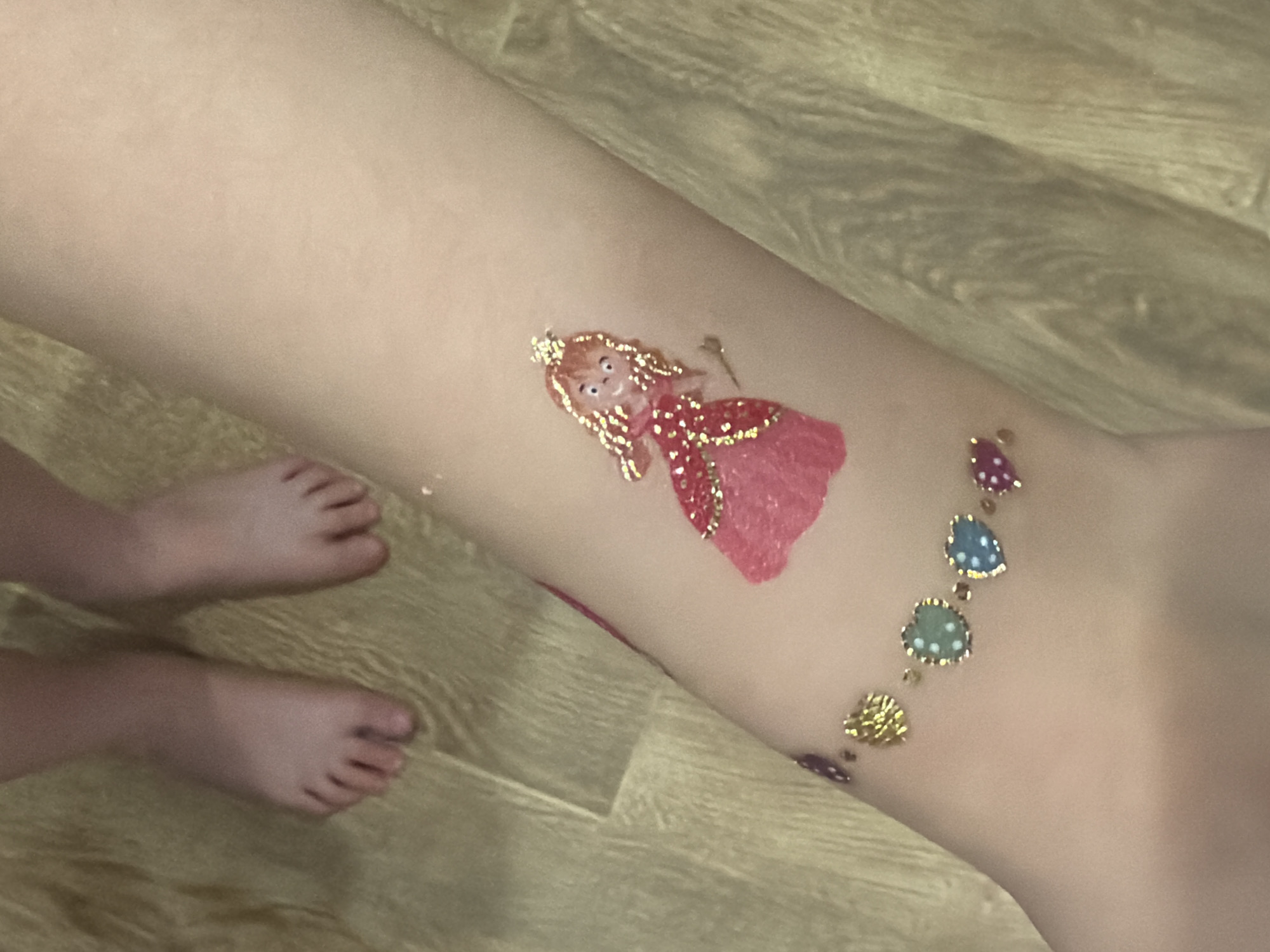 The image captures a child's lower leg and bare feet, adorned with colorful temporary tattoos, set against a light-toned wooden floor.
On the leg, a prominent temporary tattoo depicts a princess-like figure. She has golden, curly hair, a small gold crown, and a sparkly pink dress with golden accents. Her face is simple, showing black eyes and a faint smile. Further down the leg, near the ankle, is another temporary tattoo resembling an ankle bracelet. This "bracelet" is composed of a chain of small, heart-shaped charms in various colors, including blue with white polka dots, teal with white polka dots, purple, and solid gold.
The child's bare feet are visible in the lower-left portion of the frame, with toes clearly defined, resting on the wooden or laminate flooring. The setting appears to be indoors, likely within a home, as indicated by the residential-style flooring. The image focuses on the temporary body art, suggesting a moment of playful childhood expression. No text is visible in the image.
