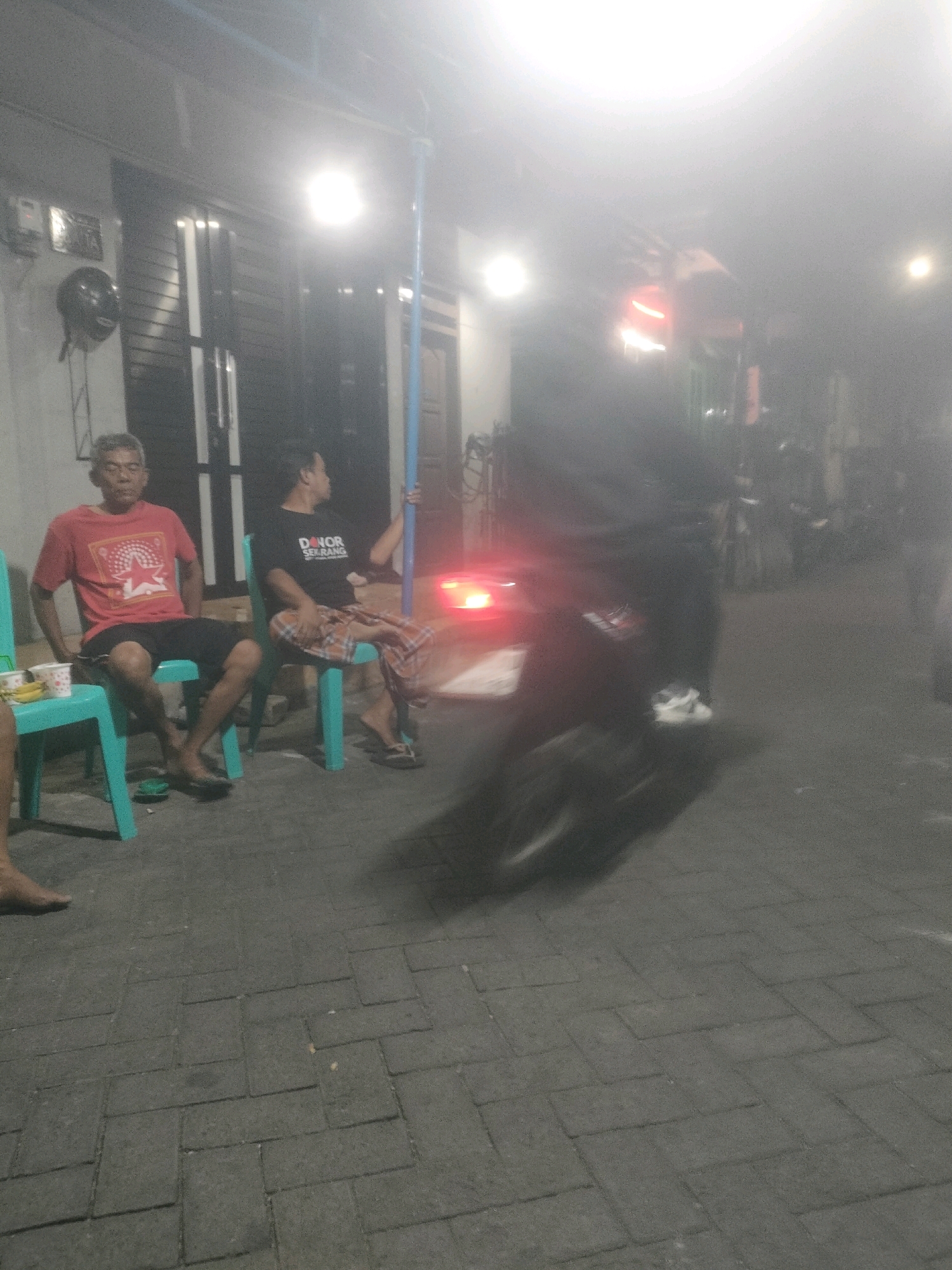 Two men are seated on teal plastic chairs on a paved sidewalk at night. The man on the left wears a red t-shirt with a graphic design and black shorts. The man on the right wears a black t-shirt with white text that reads "DONOR SEKARANG" and patterned shorts. Both men are barefoot. To the left of the man in the red shirt, there are two white cups and some food on a small white container. A motorcycle with a rider is blurred in the foreground, moving from left to right across the frame. The motorcycle's brake light is illuminated red. In the background, there is a building with a closed metal door and a bright overhead light fixture. The scene appears to be a residential or commercial street in Surabaya, Indonesia, indicated by the language on the t-shirt.