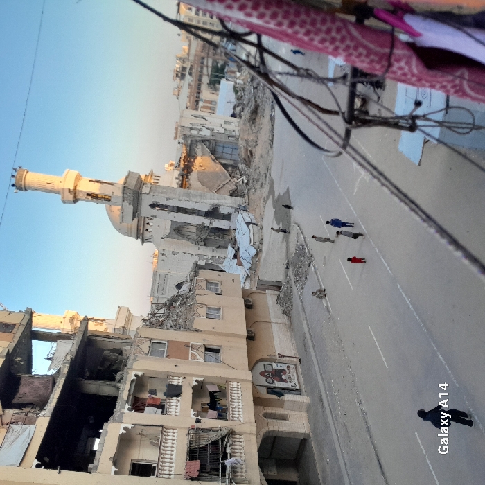 The image depicts extensive urban destruction in the city of Khan Yunis, Palestinian Territory, under a clear, light blue sky. In the background, a large mosque, identifiable by its distinct minaret and dome, is severely damaged, with its dome partially collapsed and significant rubble at its base. The minaret remains standing but shows damage. To the left, a multi-story building, likely residential, exhibits severe structural damage, with several upper floors partially collapsed and interiors exposed. Laundry hangs from balconies on the undamaged lower floors of this building. The street in the foreground is paved with visible white lane markings and is relatively clear of heavy debris, though smaller piles of rubble are scattered near the damaged structures. Approximately seven individuals are present on the street: one person in dark clothing walks away in the lower-left foreground, a group of four children or young individuals, one in a red shirt, stand near the center, one person sits on the left curb, and another is further down the street towards the mosque. Electrical wires and a patterned fabric are visible in the immediate right foreground, partially obscuring the view. The watermark "Galaxy A14" is present in the lower-left corner.