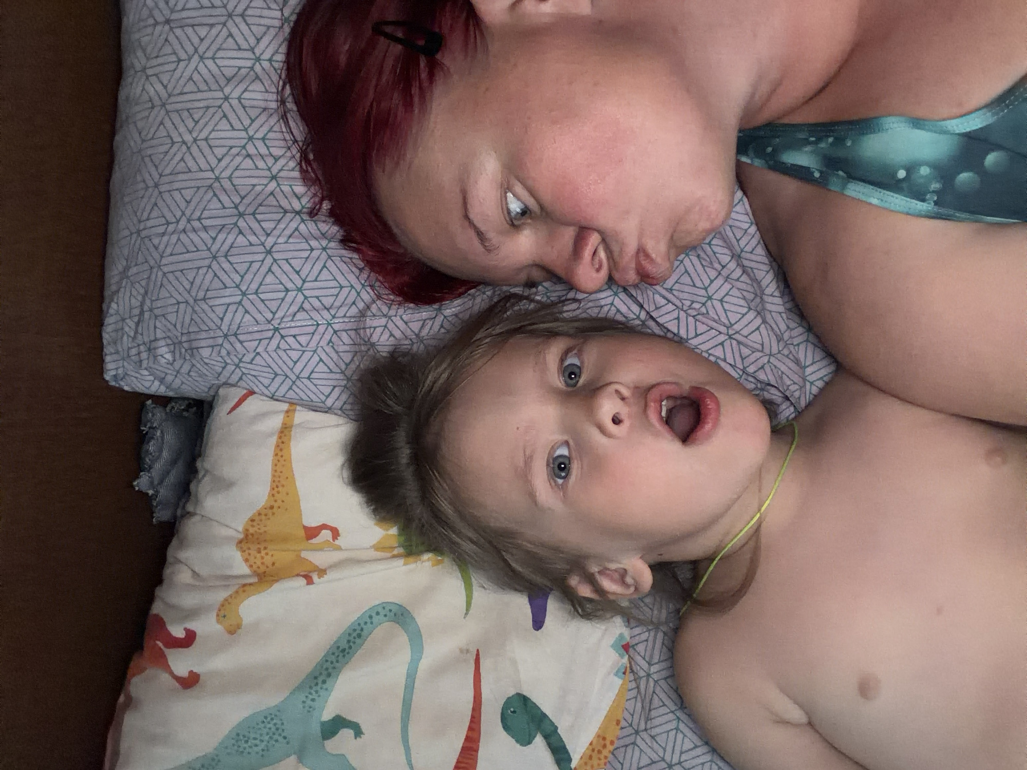 The image depicts an indoor scene in Chaikovsky, Russia, featuring two individuals, an adult female and a child, lying down side-by-side. The setting appears to be a bed, with pillows and bedding visible.
On the right, an adult female with reddish-auburn hair is positioned, her head resting on a pillow with a light-colored geometric pattern. She wears a teal-colored top with a spotted design. Her face is turned towards the child, with her lips pursed in a playful "kissing" expression. A dark hair clip is visible in her hair above her left ear.
On the left, a young child with light brown hair tied in a bun is positioned, their head resting on a white pillow adorned with colorful dinosaur illustrations. The child is shirtless and wears a thin, bright green necklace. Their mouth is wide open in a surprised or playful "O" shape, and their eyes are directed towards the camera.
Between the two individuals' heads, a crumpled piece of light blue fabric is visible. The background above the pillows is a solid, dark brown, likely a headboard or wall. The lighting suggests a dimly lit indoor environment.