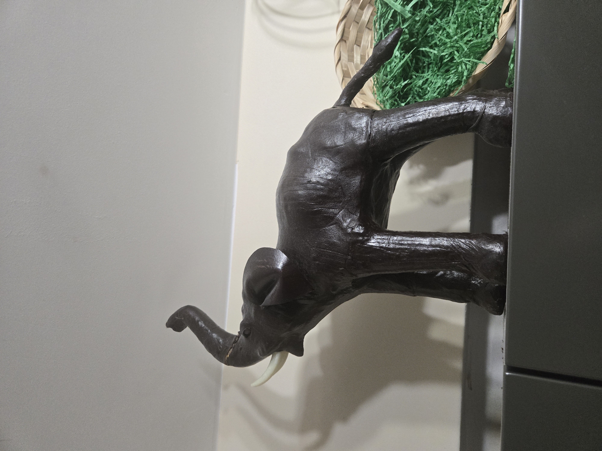 A brown, stylized elephant figurine stands in a room. The elephant is positioned such that its head and trunk face towards the left side of the image. The elephant's tusks are visible. The figure appears to be emerging from a gray object to the right side of the image, and above the elephant, there is a woven basket containing green shredded paper. The background consists of a pale wall. The location and city are unknown.
