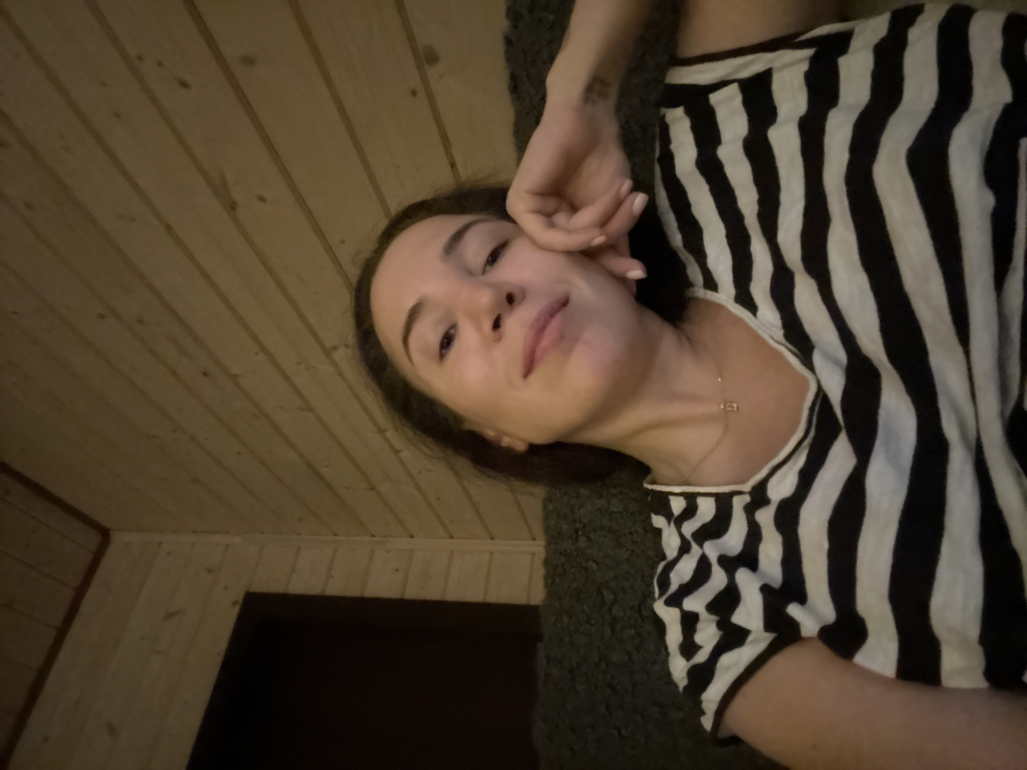 A woman is taking a selfie in a room with wood-paneled walls. She is wearing a black and white striped shirt and a necklace with a small pendant. Her head is resting on her hand. The room appears to be dimly lit. There is no discernible information in the image to indicate the location is the city of Stupino, Russia.