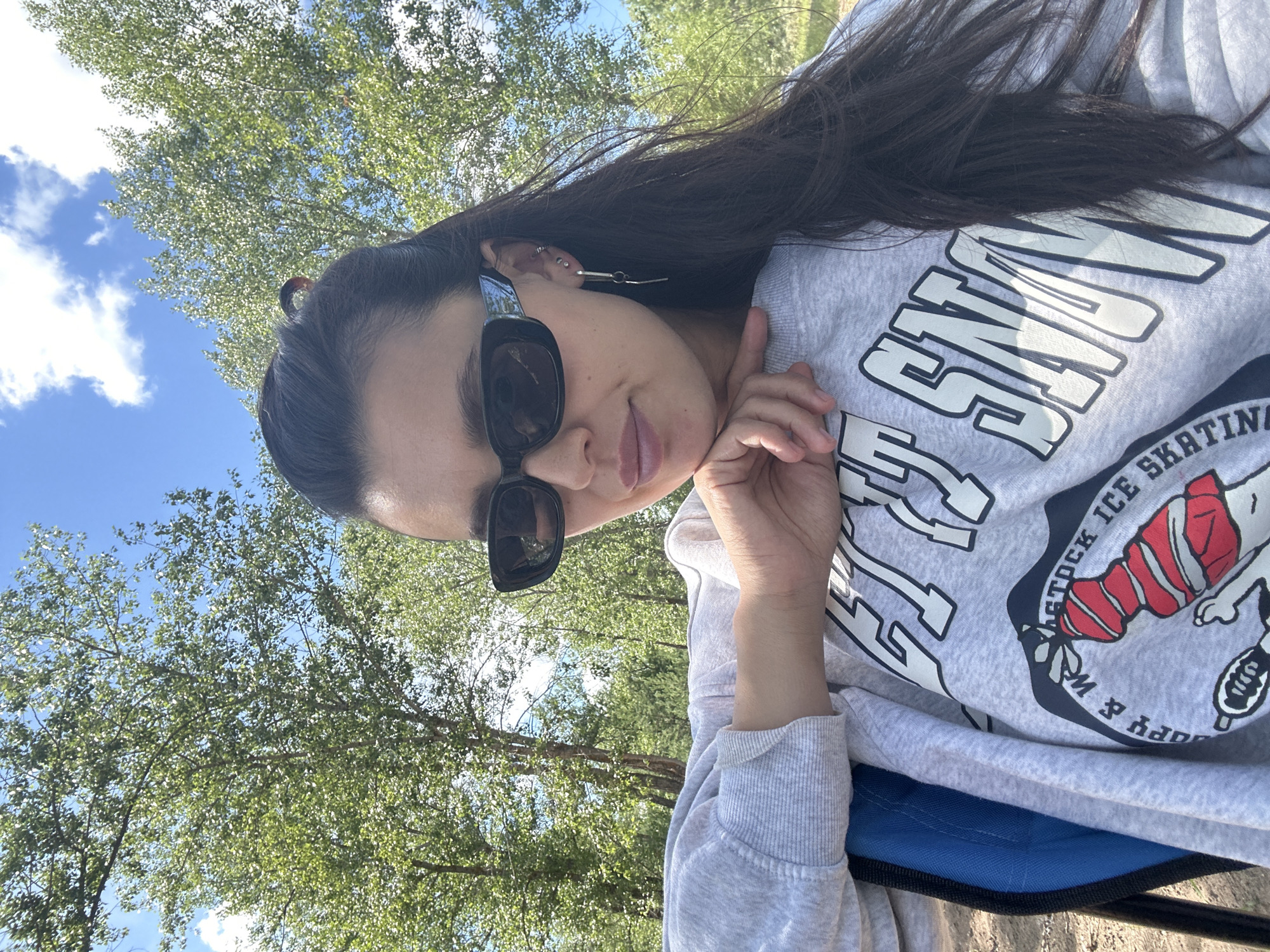 A woman with dark hair, wearing black sunglasses and a light gray sweatshirt, is sitting in a blue chair outdoors. She is resting her chin on her hand and looking towards the camera. The sweatshirt has large white lettering and a graphic of a cartoon character with red and white stripes. In the background, there are trees with green foliage and a blue sky with white clouds. The scene appears to be a park or recreational area. The location is the city of Noviy, Russia.