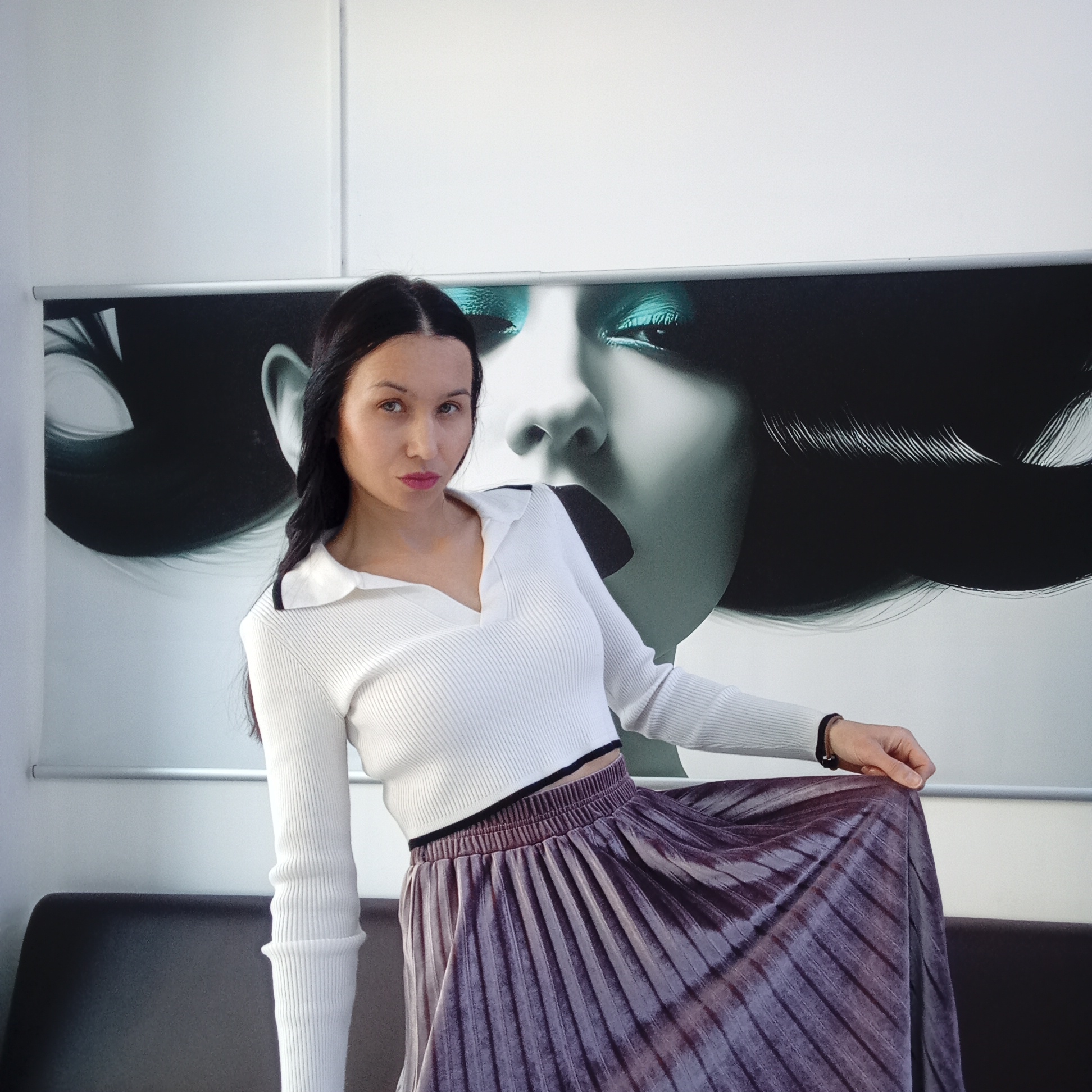 A young woman with long, dark hair parted in the middle is featured in a half-length portrait, posing indoors. She is wearing a white ribbed knit crop top with a collar and V-neck, paired with a long, pleated skirt in a shimmering dark purple or maroon hue. Her expression is neutral and direct, looking towards the camera, with a hint of pink lipstick. Her left hand is visible, gently holding a part of her skirt.
Behind her is a large, wall-mounted banner or poster featuring a stylized close-up of a woman's face. The banner's image is predominantly black and white, but the eyes of the depicted face are strikingly highlighted with bright teal eyeshadow. The hair on the banner is long and dark, flowing. The banner is positioned against a plain white wall, with a thin silver-colored bar visible along its top edge.
The woman is seated or leaning on a dark-colored, possibly leather or faux leather, couch or bench that occupies the lower section of the frame. The setting appears to be a modern indoor space such as a studio, office, or commercial establishment, suggesting a fashion or beauty context. There is no visible text in the image. The location context is Izhevsk, Russia.