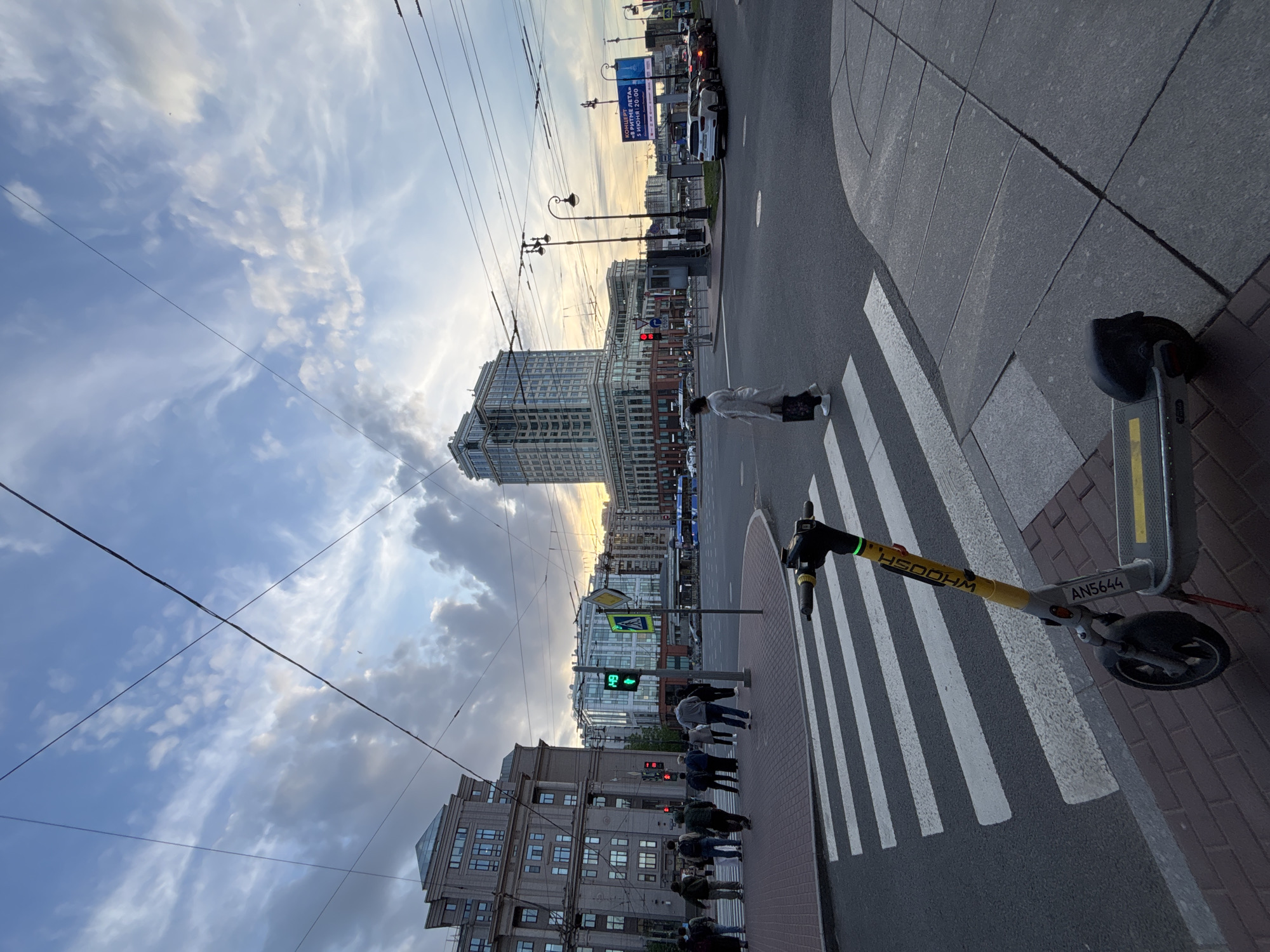 The image captures an urban street scene in Saint Petersburg, Russia, likely during the late afternoon or early evening given the low sun and warm light on the horizon. The view looks down a wide city street at an intersection with a prominent pedestrian crosswalk (zebra crossing).
In the foreground, an electric scooter, possibly a rental, is parked on the sidewalk next to the crosswalk. It is grey and yellow, with the text "HSOOHM" and "AN5644" visible on its body. Several pedestrians are visible, some waiting at the curb for the light to change, and at least one person is crossing the street. A green pedestrian traffic light is clearly visible, indicating it's safe for pedestrians to walk. A red vehicle traffic light is also visible further down the street.
The street is lined with multi-story buildings of varying architectural styles, suggesting a mix of older and modern structures. Overhead wires, characteristic of trolleybus or tram lines, crisscross the sky. On the right side of the street, a large blue billboard displays Russian text: "КОНЦЕРТ", "РИТМЕ ЛЕТА!", "5 ИЮНЯ | 20:00" (Concert, Rhythm of Summer!, June 5 | 20:00). Cars are visible on the street, some parked and others in traffic.
The sky is partly cloudy with a mix of wispy cirrus clouds and more substantial cumulus clouds, creating a dynamic backdrop. The overall impression is one of a bustling city environment during the transitional hours of the day.