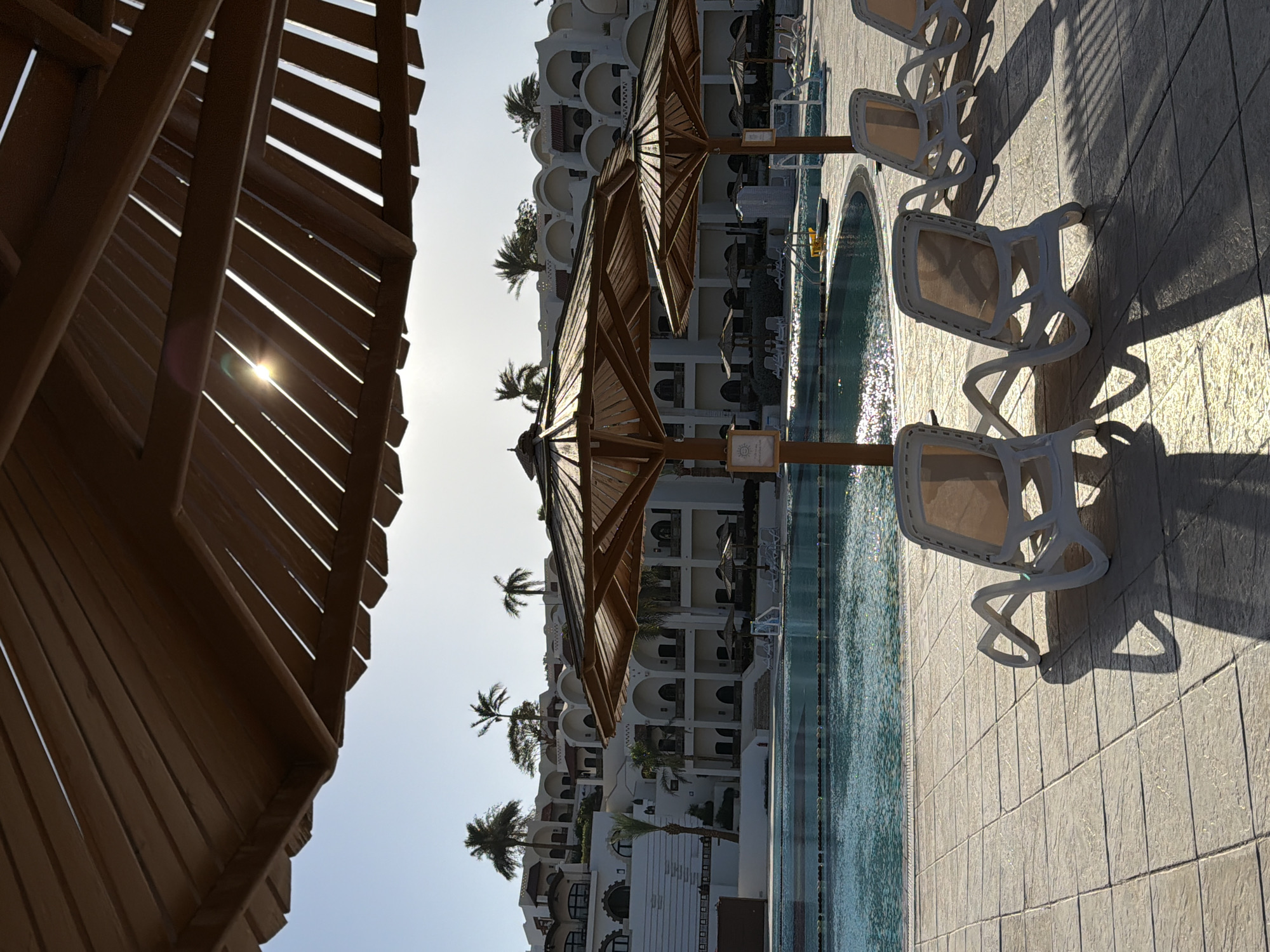 This image captures a vibrant outdoor resort scene in Sharm El Sheikh, Egypt, looking towards a hotel and swimming pool, with the sun low in the sky.
In the foreground on the left, the dark, slatted wooden underside of a large pergola or umbrella frames the shot. Sunlight filters through the gaps, creating a prominent lens flare on the left. Moving into the midground, a pristine blue swimming pool stretches across the frame. Along its edge, several wooden umbrellas with slatted tops are neatly arranged, casting long shadows onto the light-colored tiled pavement. Adjacent to these umbrellas are multiple white and beige sun loungers, also casting extended, defined shadows that indicate either early morning or late afternoon.
The background features a multi-story hotel building, characterized by its light-colored facade and numerous arched windows and balconies, typical of regional architecture. Several slender palm trees are visible in front of and around the hotel, adding to the tropical resort atmosphere. The sky is clear and bright, consistent with sunny weather.
The scene suggests a peaceful, relaxing holiday environment, devoid of people, which emphasizes the tranquility. The sparkling pool water and strong shadows contribute to a sense of warmth and an inviting setting. A small, sun-like emblem is visible on a sign attached to one of the umbrella poles, but no clear text can be discerned. The overall impression is one of an idyllic, sun-drenched resort ready for guests to enjoy.