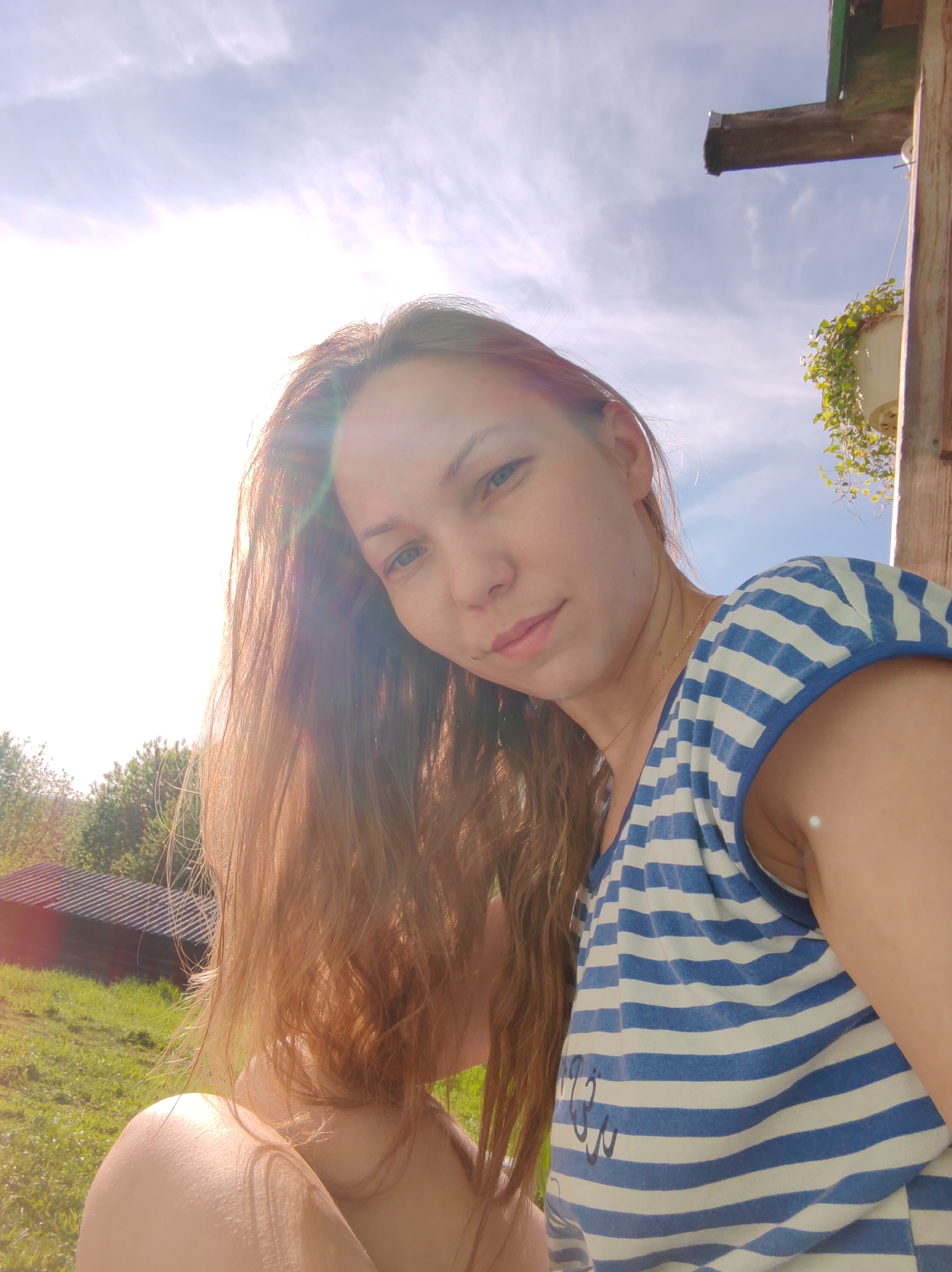 A woman with long brown hair and fair skin is looking at the camera. She is wearing a blue and white striped shirt. The background includes a building with a dark roof, green grass, trees, a blue sky with white clouds, and a wooden structure with a hanging plant. The location may be Чухлэм, Russia.