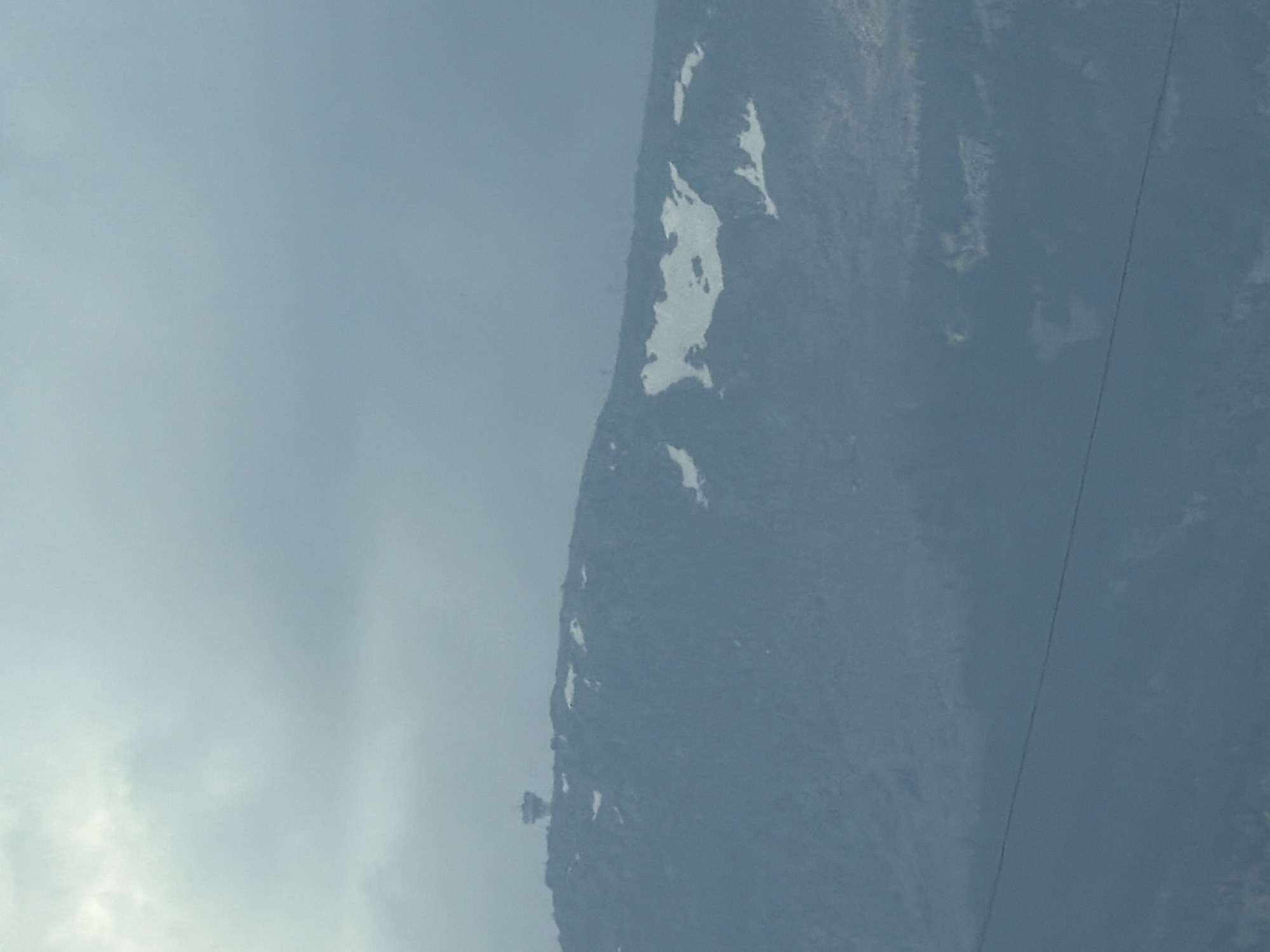 The image shows a wide shot of the Vitosha Mountain near Sofia, Bulgaria. A structure, likely a tower or building, is visible on the summit of the mountain. Patches of snow are scattered across the mountainside, suggesting a cold or high-altitude environment. The sky is overcast with gray clouds. In the foreground, a thin, dark line extends horizontally across the frame, possibly a power line or cable. The overall scene is muted and appears to be captured in low light conditions.