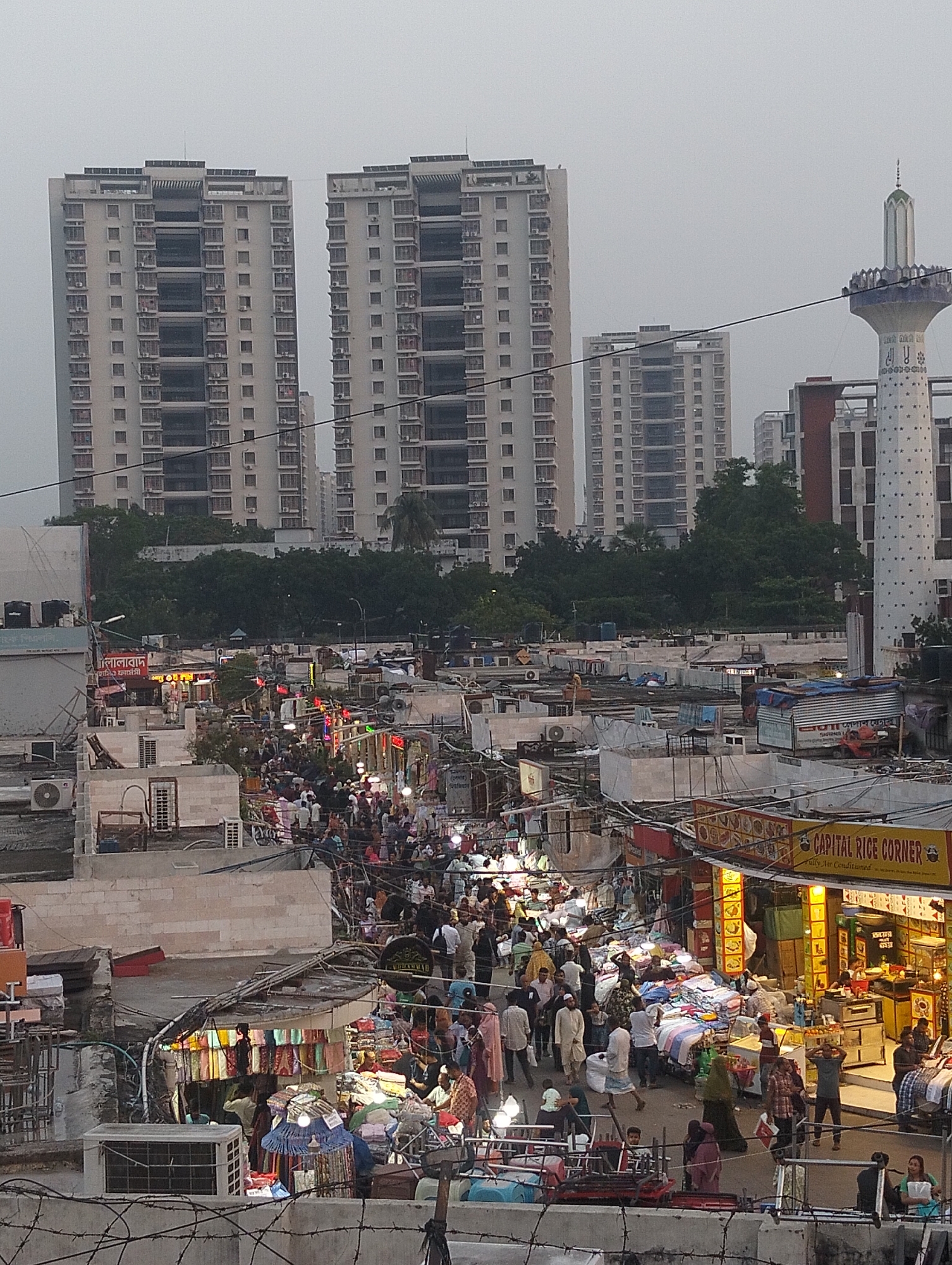 The image shows a crowded marketplace in Dhaka, Bangladesh. The scene is dominated by densely packed stalls and shops lining narrow streets, filled with people shopping and walking. The shops appear to be selling textiles, food, and various consumer goods. The "CAPITAL RICE CORNER" stall is visible. The architecture is a mix of low-rise concrete buildings and structures with corrugated metal roofs. In the background, modern high-rise apartment buildings and a minaret with white and blue decoration are visible above the lower rooftops of the commercial area. Barbed wire fencing is visible in the foreground. The lighting indicates either late afternoon or early evening. The place is likely located in the old part of Dhaka.