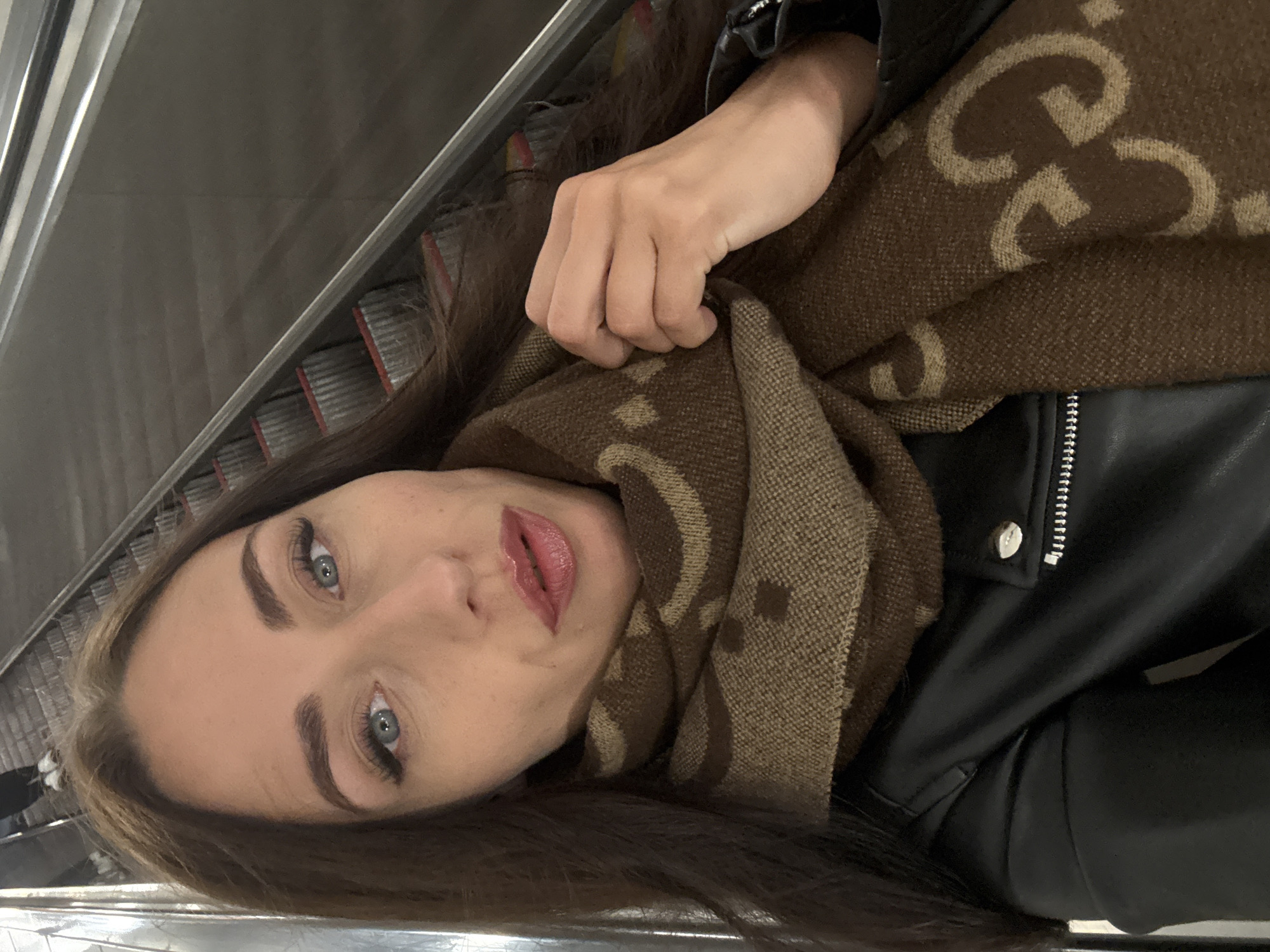 The image is a selfie taken by a woman with long, dark brown hair and blue eyes. She is wearing a black leather jacket and a brown scarf with a repeating pattern of interlocking "G" shapes. Her left hand is holding the scarf near her neck. Behind her, an escalator with red-edged steps is visible, suggesting she is in a public transportation hub or a shopping center. The lighting is dim and appears to be artificial. There is no specific identifiable landmark of Saint Petersburg visible in the image. The woman's makeup is subtle, with emphasis on her eyes and lips. The overall impression is of a casual, everyday moment captured on camera.