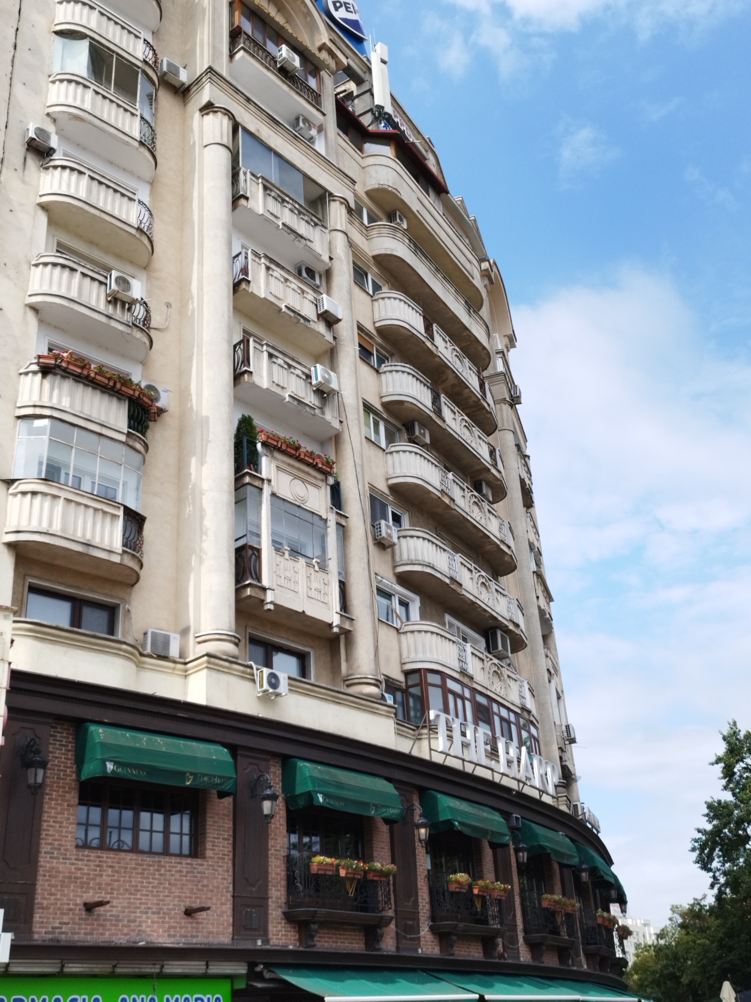 The image shows a side view of an apartment building in Bucharest, Romania, under a partly cloudy blue sky. The building is tall and has a light-colored facade with several floors, each featuring curved balconies with ornate railings. Many air conditioning units are visible on the balconies and walls.
The lower section of the building is a commercial space with dark brick walls and green awnings. The awnings are branded with the word "GUINNESS" and some text that appears to read "THE HARP" on the right side of the image. Beneath the awnings, there are windows and decorative ironwork balconies with flower pots. In the foreground, a green awning with white text is partially visible at the bottom. Trees and other buildings can be seen in the background, suggesting an urban environment.