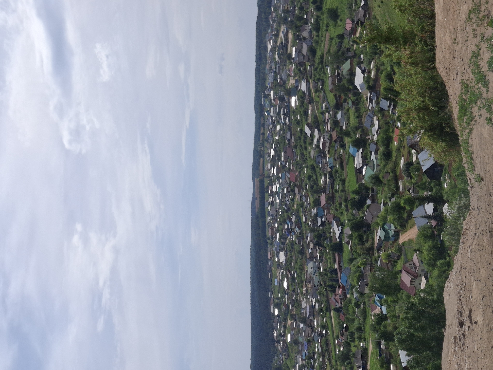 A sideways view of a village landscape with a cloud-filled sky on the left and a densely populated area on the right. The village contains numerous buildings with varying roof colors intermingled with green trees. The rightmost edge of the image features an earthen formation covered with sparse vegetation. The buildings and foliage hint at the location being in a rural or suburban part of Ольховка, Russia, but without specific landmarks, precise identification within the city is challenging.