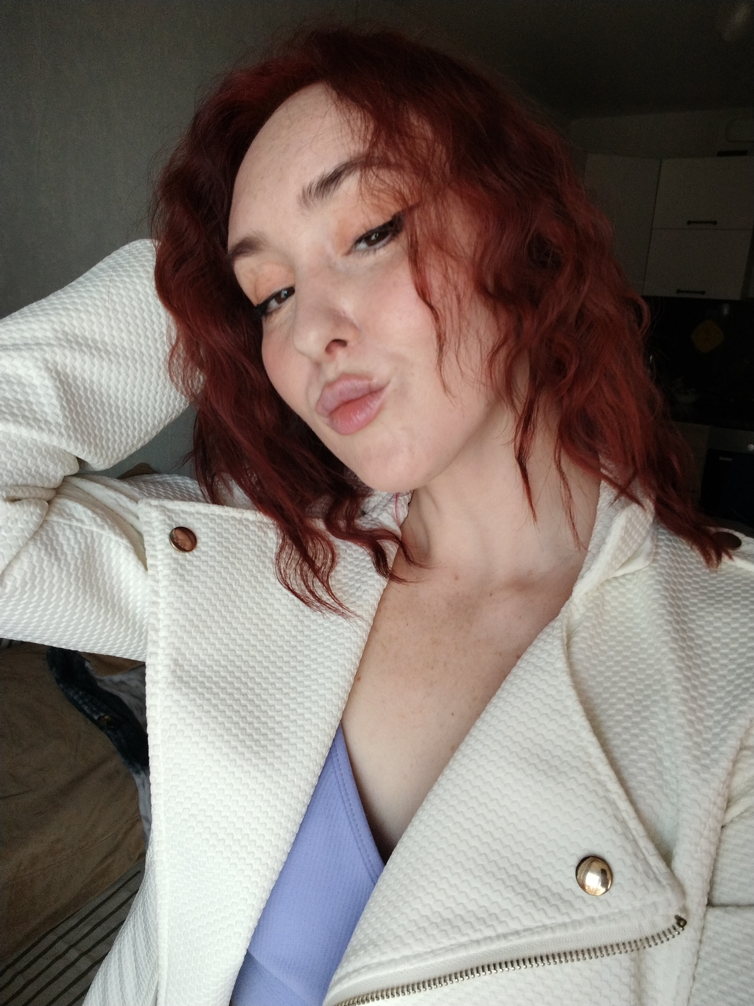 A woman with red curly hair is posing for a selfie indoors. She is wearing a white textured jacket over a light blue top. Her left hand is behind her head. The background includes a light gray wall and kitchen cabinets. The location in the city of Izhevsk, Russia, is not discernable from the image.