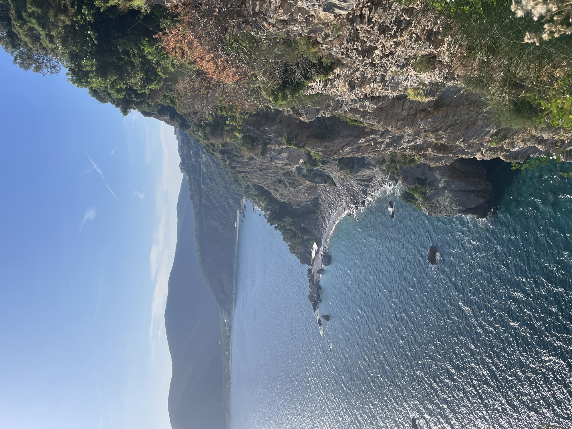 The image depicts a coastline view near Budva, Montenegro. The camera is positioned on a cliff overlooking the Adriatic Sea. To the left is a distant mountain, and the sea reflects sunlight, creating a shimmering effect. The cliffside vegetation consists of trees and some arid growth. The sea is a mix of turquoise and dark blue. There are no people visible in the frame.