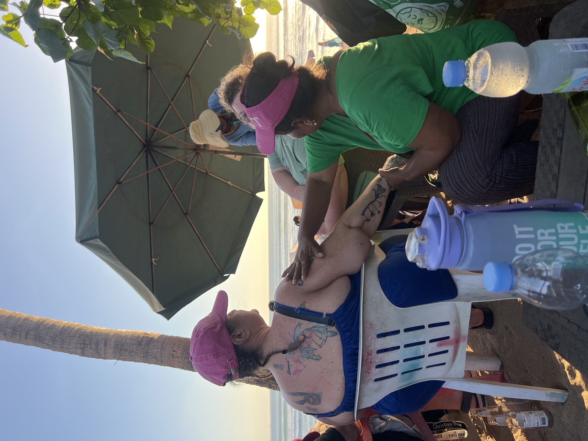 In Kuta, Indonesia, at sunset, four individuals are gathered near the beach. A woman in a blue dress and pink cap, facing away from the camera, is seated on a white plastic chair. Her back is adorned with tattoos, and she is extending her left arm, which is being touched by another person. To her right, a woman in a green shirt and pink visor is kneeling and appears to be speaking with the first woman. Behind them, a person wearing a light-colored sun hat is visible under a large green umbrella. The setting includes a sandy beach, a palm tree, and the ocean in the background. On a wooden table in the foreground, several water bottles and an empty beer bottle are present.