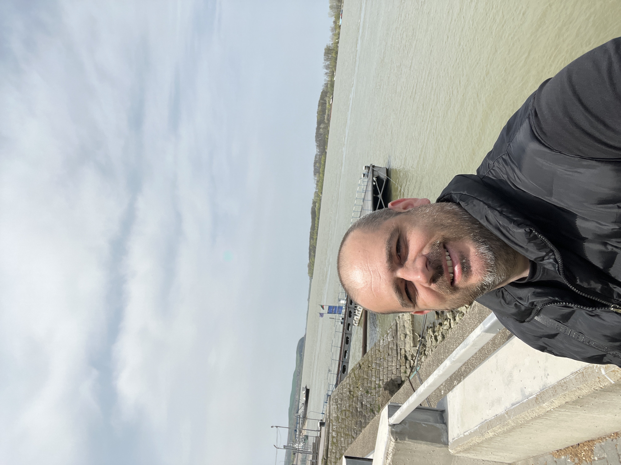 A selfie of a smiling man with a shaved head and a dark jacket. Behind him is a river with a platform dock. The background also includes trees along the far side of the river. The sky is overcast. This image may be taken at the Nikopol, Bulgaria riverfront, based on the river and dock features.