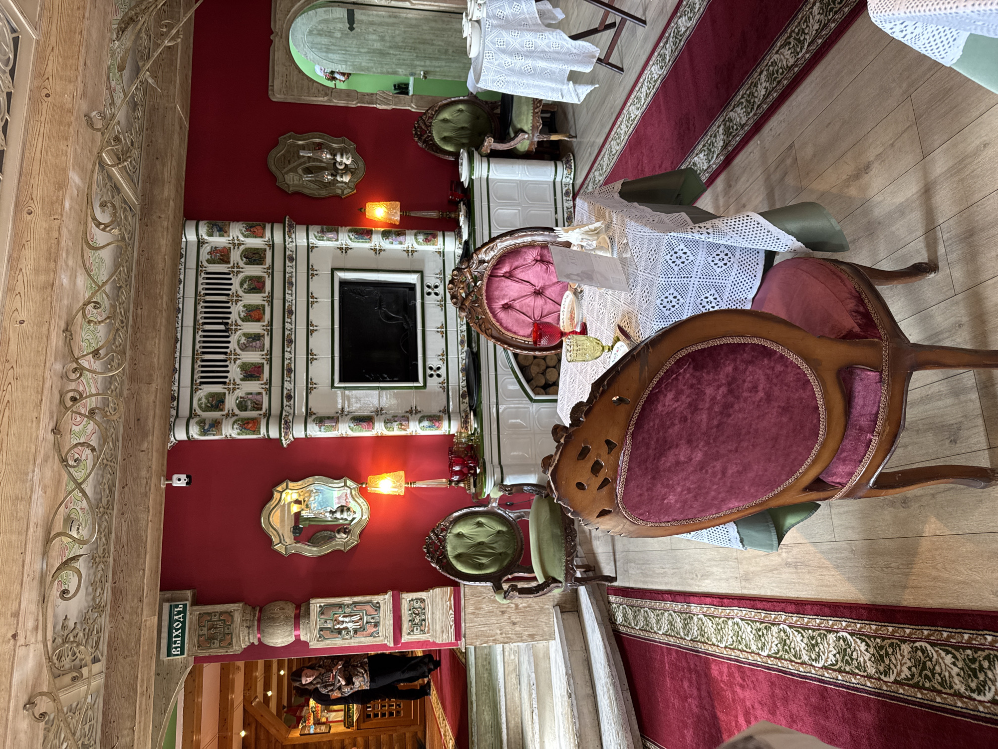 The image depicts the interior of a restaurant with a rustic, traditional aesthetic. The dominant color is a deep red, featured on the walls and carpeting. In the center of the scene, a tiled stove with ornate paintings is a prominent feature, with a black television screen embedded within it. Two tables are set for dining; one in the foreground is covered with a white lace tablecloth and has velvet chairs, while a second table to the right is also covered with a white tablecloth. Several chairs, some upholstered in green velvet and others in dark wood with burgundy upholstery, are placed around the tables. A decorative mirror hangs on the red wall above a lamp with a yellow shade. In the background, a staircase leads up to an area where two individuals are standing near a wooden structure. A green door is visible on the far right side of the image. The ceiling is made of exposed wooden beams, and decorative metalwork is attached to the ceiling. An "EXIT" sign is visible on the upper left. The lighting is warm, with lamps casting a reddish glow.