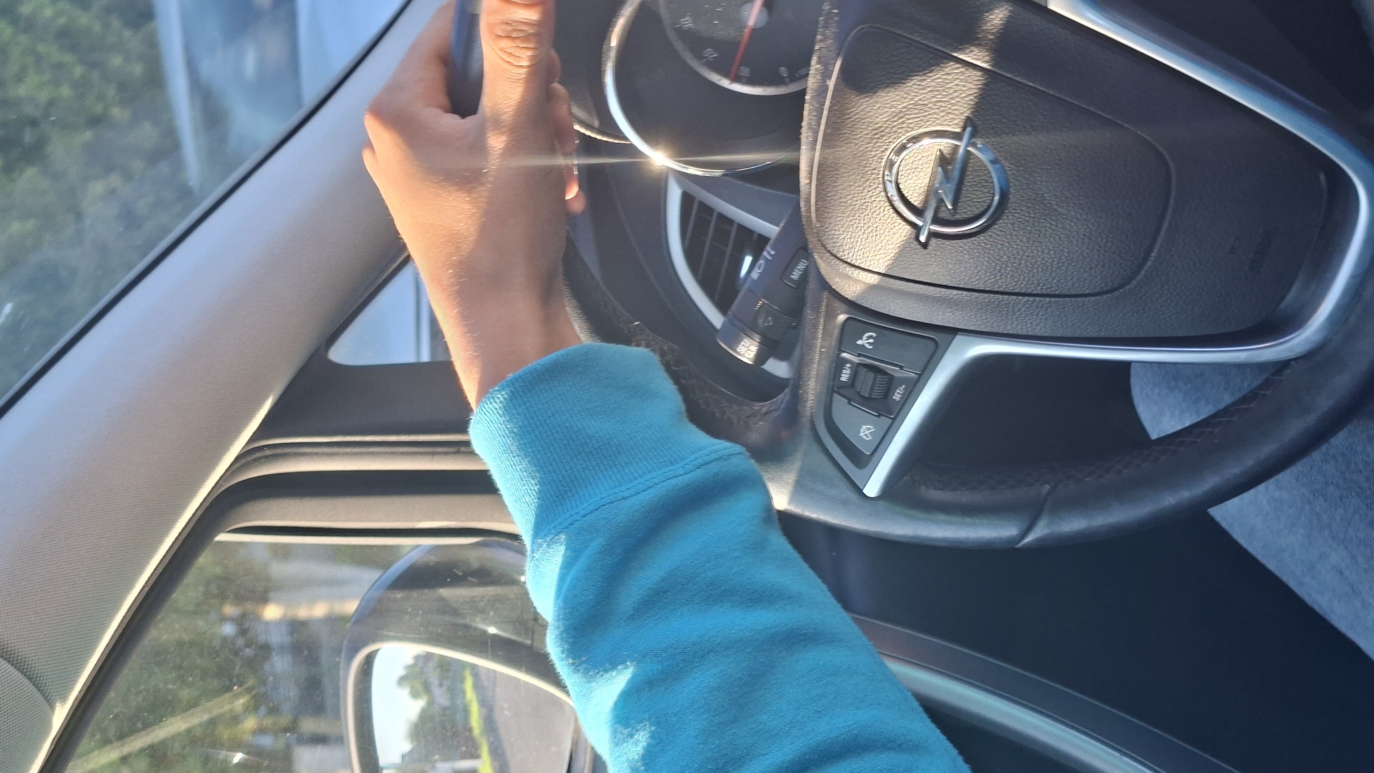 An exterior view of a person's right arm and hand on a car's steering wheel is captured from a side angle. The person is wearing a bright blue long-sleeved sweatshirt. The steering wheel, which has an Opel logo in the center, features various buttons and controls, including cruise control options. The car's dashboard, air vents, and a side mirror are also visible. Sunlight illuminates the scene, creating glare on the dashboard and steering wheel. Outside the car, a blurred background shows trees and houses, suggesting a residential or suburban area. The location is the city of Smolevichi, Belarus.