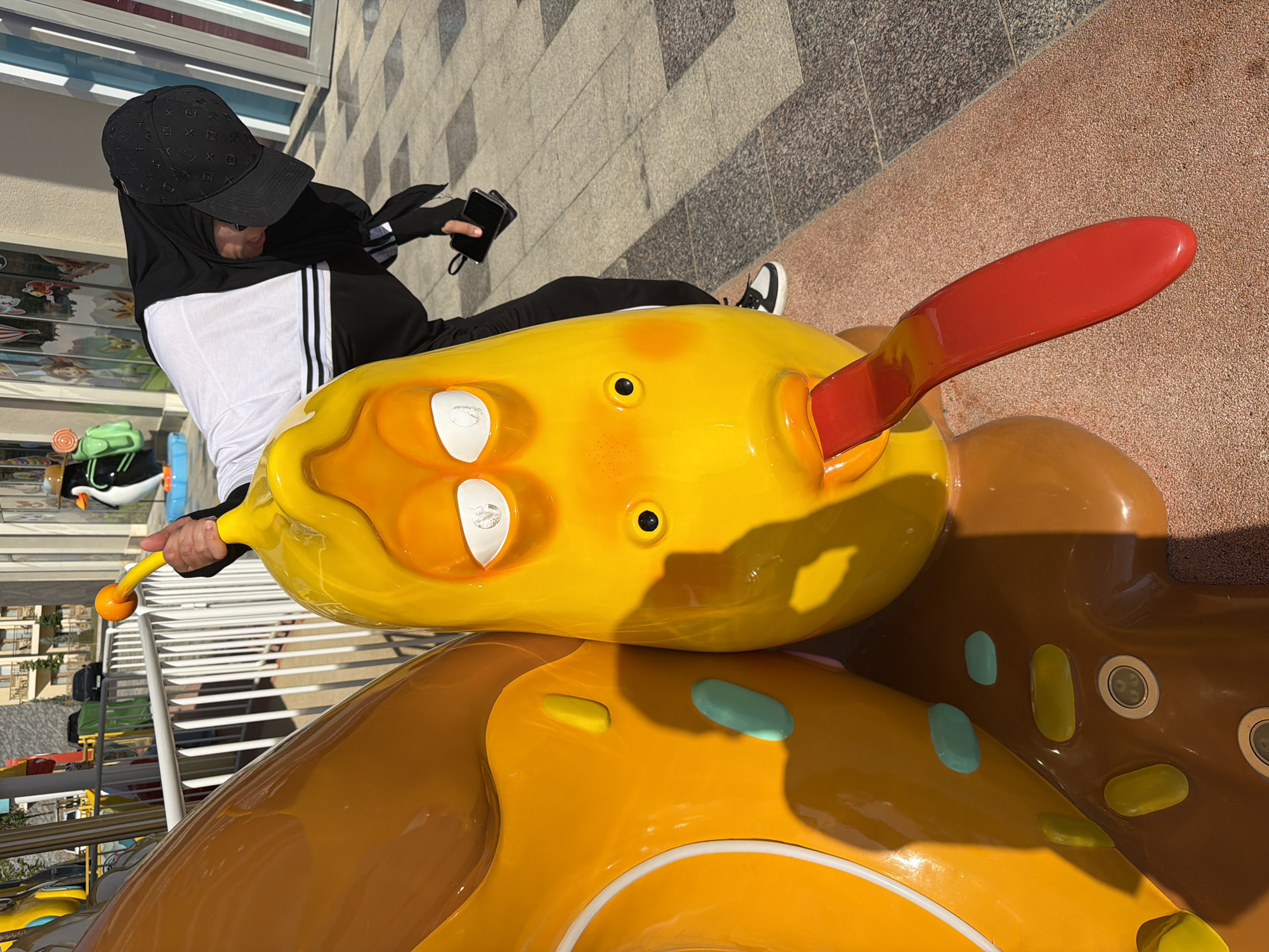In Chinarkent, Uzbekistan, a person wearing a black hijab and a white shirt with black stripes is interacting with a large, yellow, cartoonish statue of a larva with a long, red tongue. The statue appears to be part of a playground or outdoor attraction. The person is holding a yellow handle connected to the larva's head, and their left hand is holding a black smartphone. The background shows buildings, fencing, and a paved ground surface. The lighting suggests it is daytime.