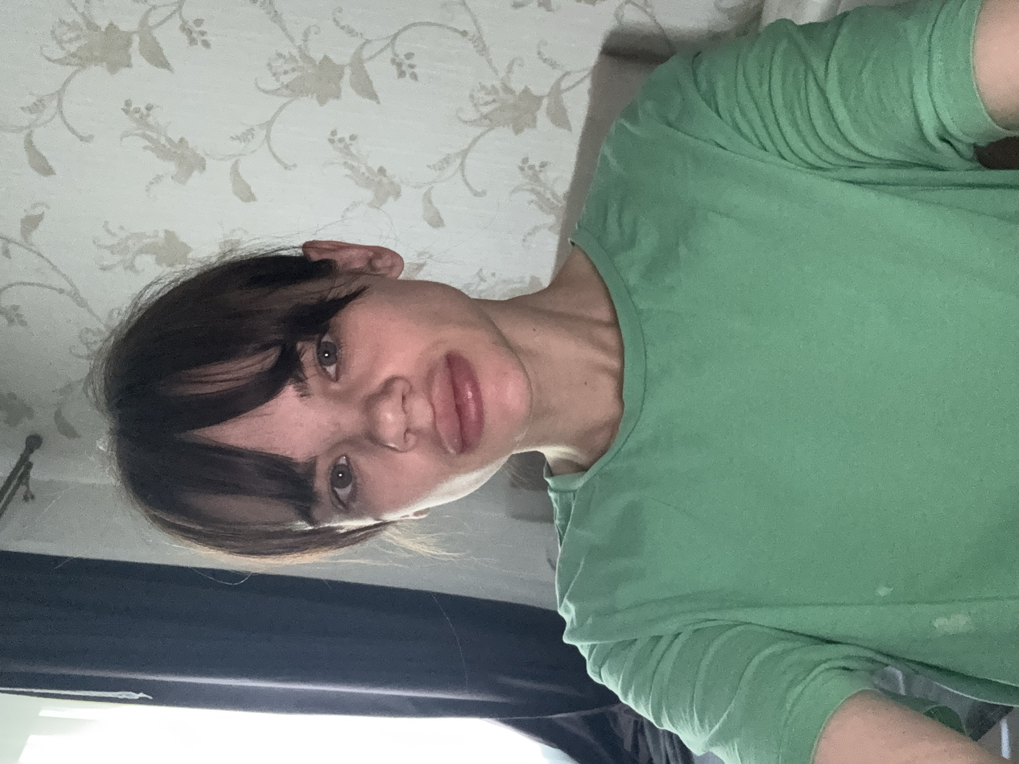 A woman with dark brown hair and bangs, wearing a green T-shirt, is indoors. The background consists of a wall with floral wallpaper and part of a window covered with a black curtain. It is not possible to determine if the woman is in Rostov-on-Don based on visual information provided.