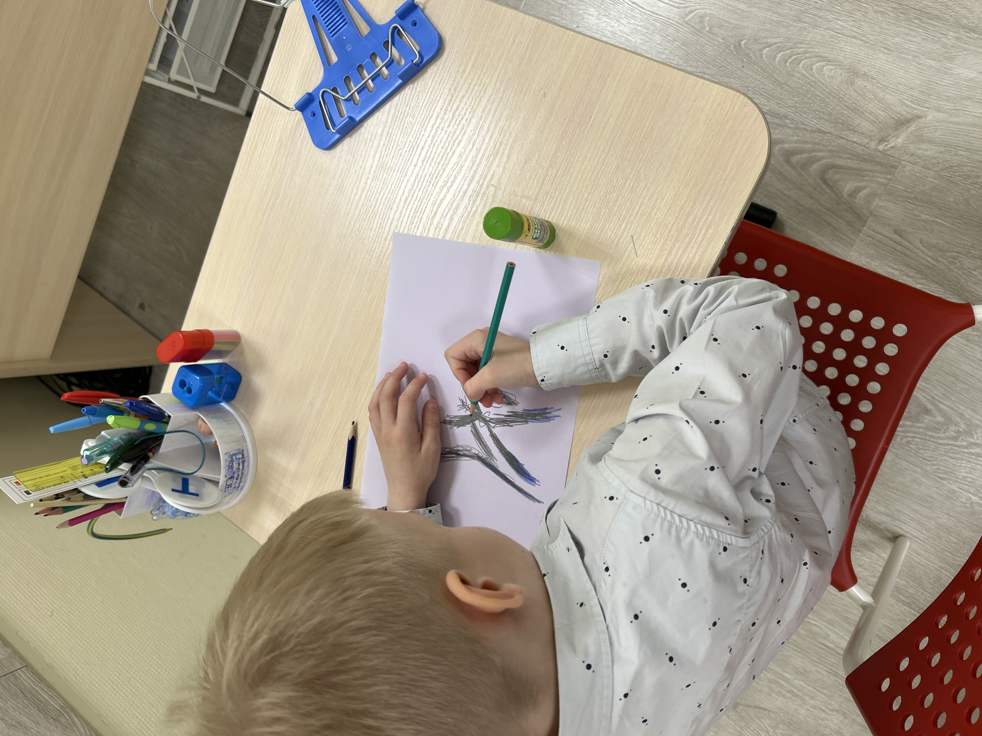 A child sits at a light-colored wooden desk drawing on a white piece of paper with a green pencil. The child is wearing a light-colored shirt with small black dots. On the desk, there is a blue tool, a container holding pens and pencils, a green glue stick, and a dark blue pencil. The child is sitting in a red chair with circular perforations. The floor is a light gray laminate. This image appears to be taken indoors, potentially in a home or educational setting. There is nothing in the image to indicate it is in Saint Petersburg, Russia.