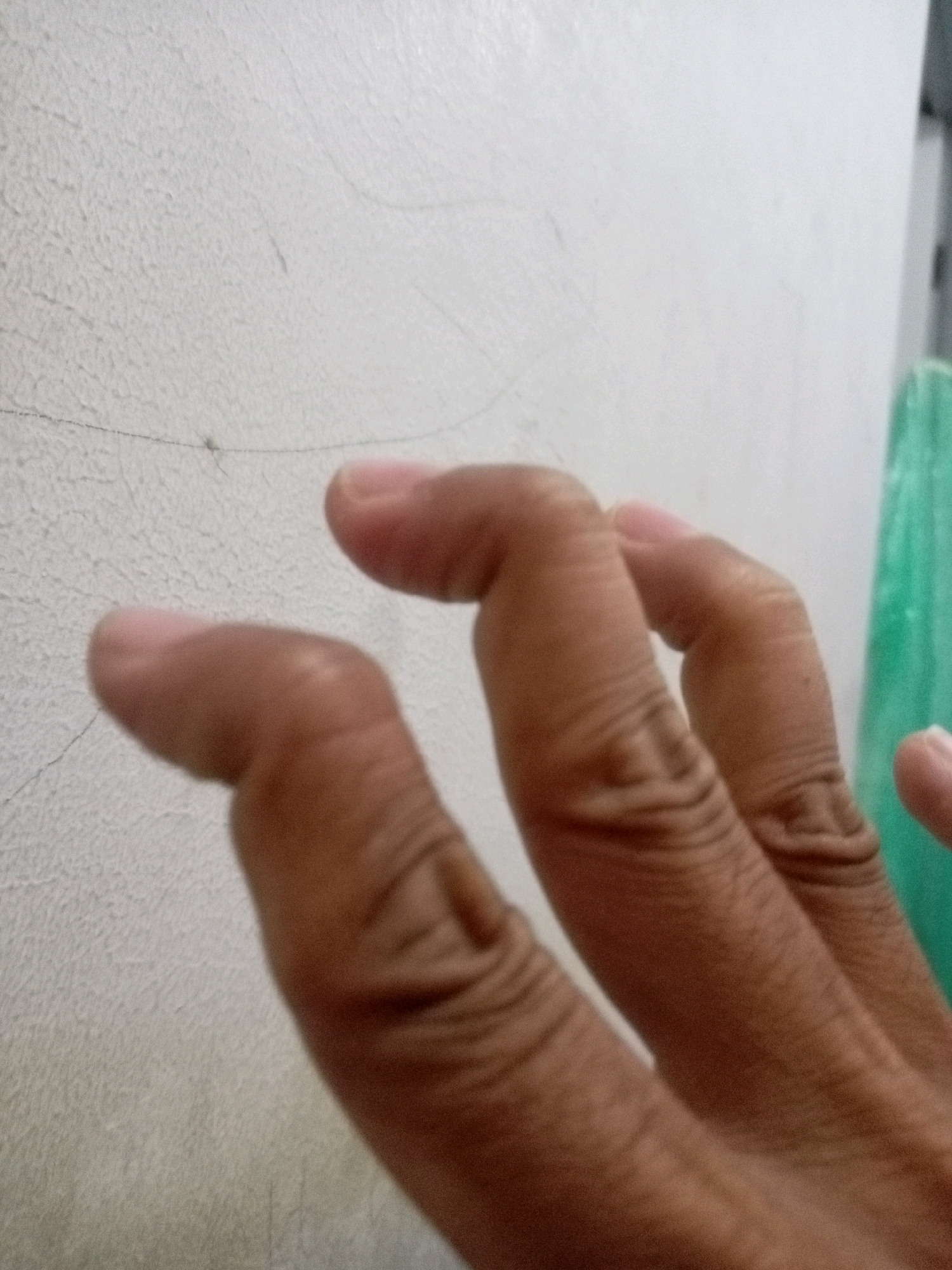 The image shows a close-up view of a person's hand, with fingers bent at unusual angles, positioned against a textured off-white wall. The wall appears to have faint linear markings and some subtle discoloration. A thin, dark strand, possibly a hair or fiber, is visible on the wall. To the right, a portion of a bright green object, possibly plastic or fabric, is visible. The lighting suggests an indoor environment, though the specific location within Batam, Indonesia, cannot be determined from the visual information provided. The focus is on the texture of the wall and the unusual posture of the fingers.